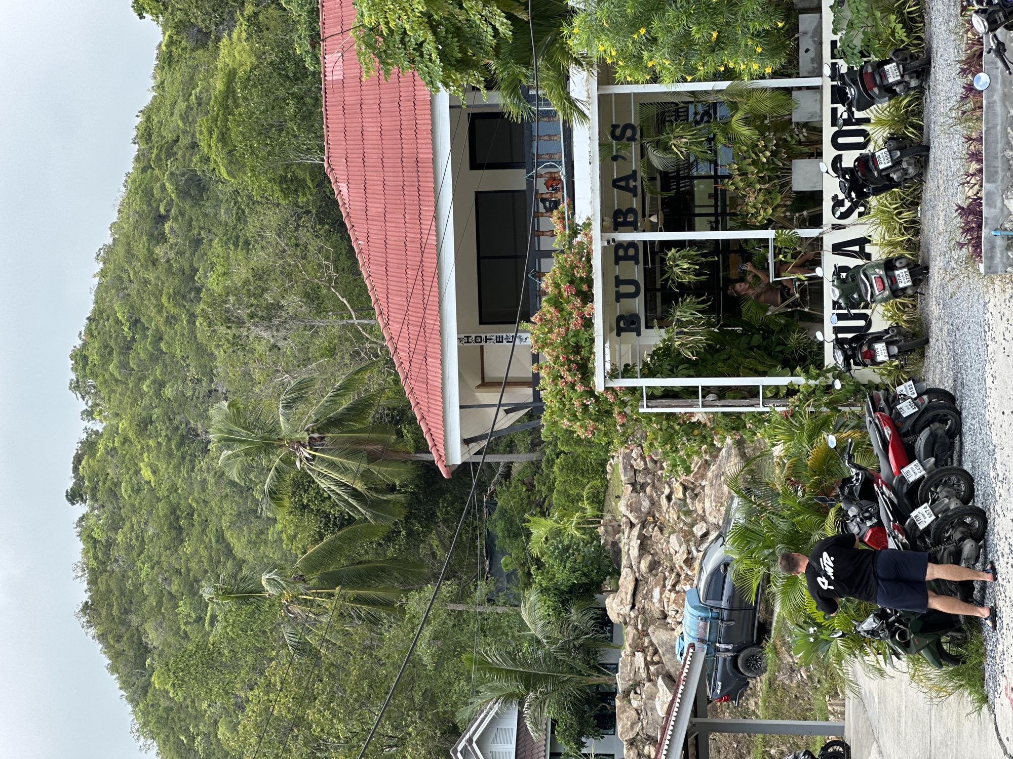 In ตำบลเกาะพะงัน, Thailand, a man wearing a black t-shirt and navy shorts stands with his back to the camera, facing a row of parked motorcycles. The motorcycles are parked on a gravel surface in front of a building with a sign that reads "BUBBA'S COFFEE". A dark-colored pickup truck with a blue tank is parked on an elevated rocky incline behind the motorcycles. Behind the coffee shop, a two-story building with a red tiled roof is visible, with palm trees and lush green foliage surrounding it. The background is dominated by a densely vegetated hillside. The sky is overcast.