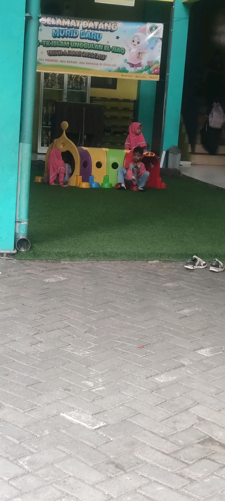 In Banjarsari, Indonesia, a vibrant scene unfolds outside a school. A colorful banner, featuring cartoon characters and Indonesian text that welcomes new students and the school year 2026/2027, hangs prominently. Below, a playground structure designed like a caterpillar, in shades of yellow, purple, and red, sits on artificial green grass.
Two young children are engaged with the playground. A girl in a pink hijab and matching outfit is partially inside the yellow section of the caterpillar structure. Beside her, a boy, also in a pink shirt and blue pants, sits on the grass, looking towards the camera. A third child, wearing a pink hijab, stands near the entrance of the caterpillar.
In the foreground, a textured gray brick pathway leads away from the play area. Two pairs of sandals are casually placed on the pathway, suggesting they were recently removed. The overall setting appears to be an outdoor educational environment, likely part of a kindergarten or elementary school, given the playful elements and the welcoming banner.