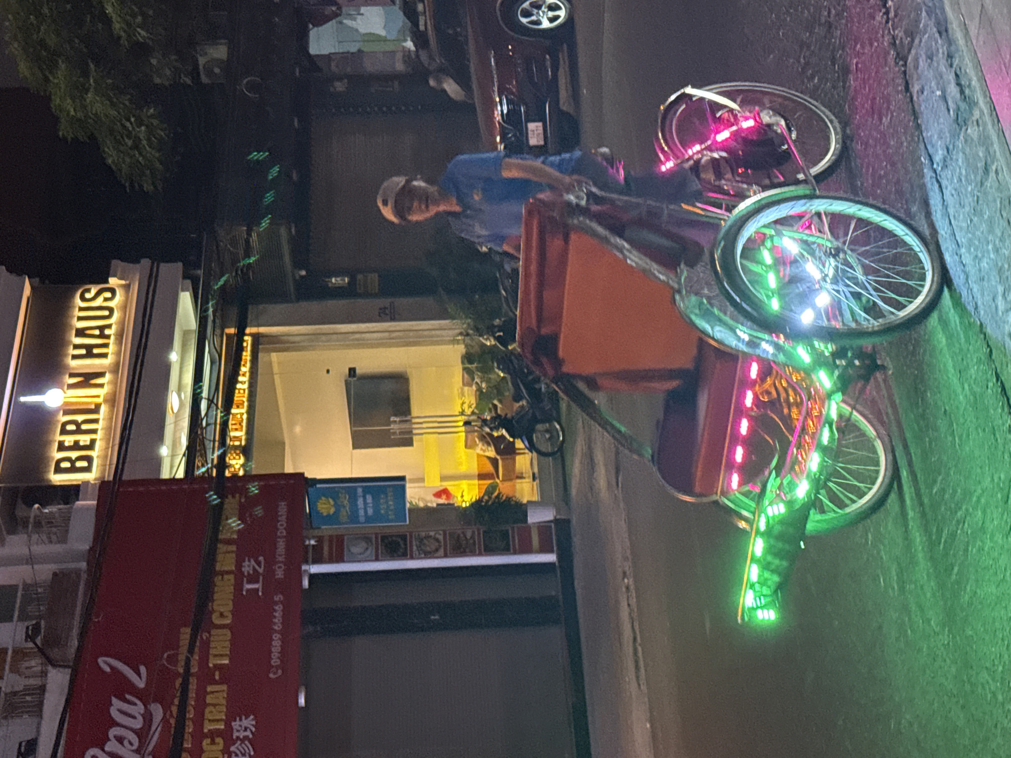 A nighttime scene in Nha Trang Ward, Vietnam, features a man in a blue shirt and cap riding a three-wheeled vehicle adorned with pink and green LED lights. The vehicle has an orange box mounted on its rear. The backdrop includes the illuminated "BERLIN HAUS" sign and a red advertising sign with Vietnamese and Chinese characters.