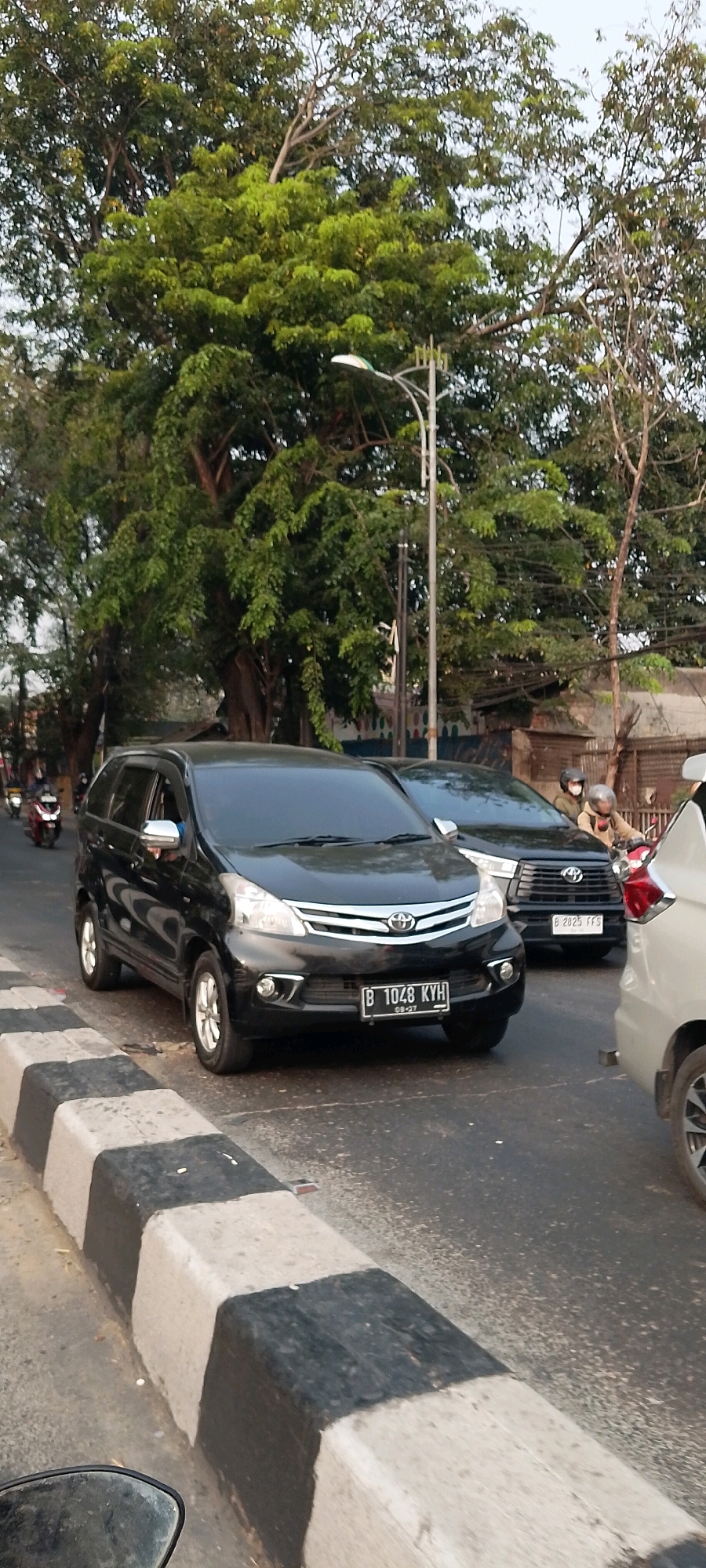 In Bekasi, Indonesia, a black Toyota Avanza, license plate B 1048 KYH, is stopped at a traffic light. The vehicle's driver is visible through the open window. Behind it, another black vehicle, possibly an SUV, also waits. Two individuals wearing helmets are visible on a motorcycle in the background, as is a light-colored car to the far right. The scene is set against a backdrop of lush green trees and a streetlight.