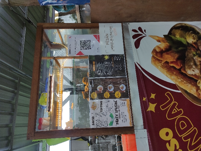 This image captures a vibrant street food stall, likely in Kecamatan Pandaan, Indonesia, during daytime. The central subject is a wooden-framed display case with glass panels, illuminated from within by a string of warm LED lights. Inside, various fried food items, possibly fritters or snacks, are showcased for sale.
To the right of the display case, a large banner with a maroon background and yellow text prominently displays "SO NDAL" (likely part of "Bakso Ndal"), alongside an appetizing graphic of a bowl of food or a wrap. Several laminated menus and signs are affixed to the back of the display case. One notable sign features a QR code for digital payments and the text "PESAN LANGSUNG BAYAR," meaning "Order, Pay Directly."
Other visible menu items include "BAKSO" (meatballs), "TOPPING," and "PAKET SPESIAL" (Special Package), with one option listed as "BRESO + ES TEH" (Breso + Ice Tea). The background on the left shows a corrugated metal wall or roof, characteristic of an outdoor or semi-permanent stall. Through the glass panels, distant trees and foliage are visible, indicating an open-air setting. No people are present, but the setup clearly indicates a ready-to-operate food vendor. The overall scene suggests a casual and accessible dining option.