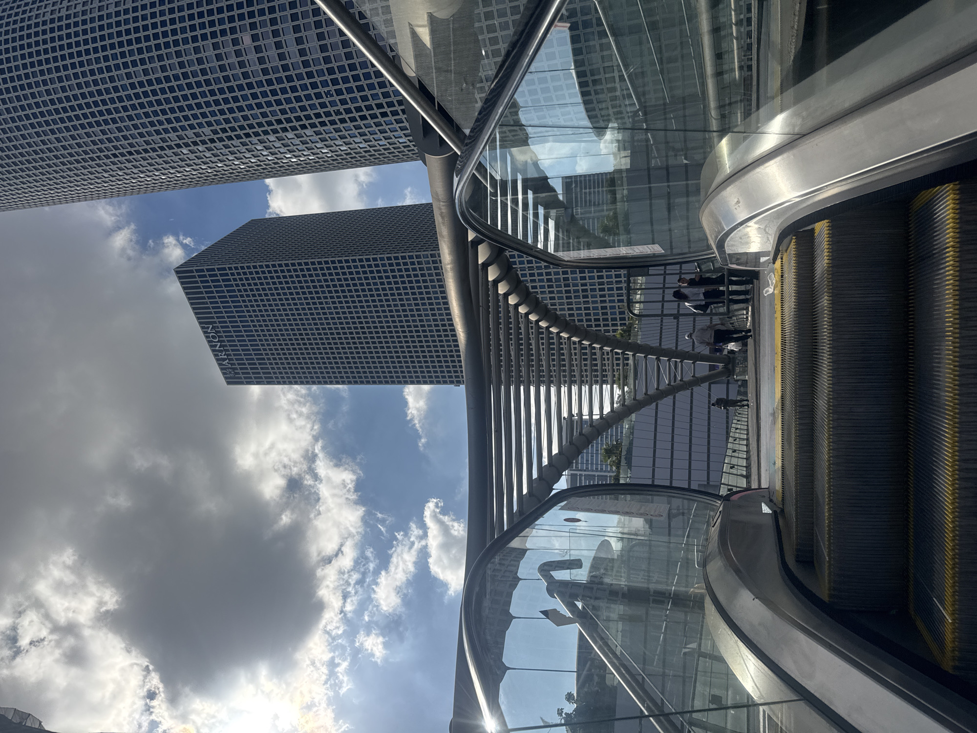 The image captures an upward view of an escalator in Tel Aviv, Israel, leading towards two modern skyscrapers. The escalator is made of metal and glass. Sunlight reflects off the glass panels, obscuring some details within. Several people are visible at the top of the escalator, some standing and talking, while others walk away. In the background, the two prominent skyscrapers dominate the frame, their grid-like facades suggesting an office or residential complex. The sky above is partly cloudy, with a mix of bright sunshine and grey clouds, creating a dynamic atmosphere. The overall scene depicts an urban environment with a blend of contemporary architecture and natural elements.