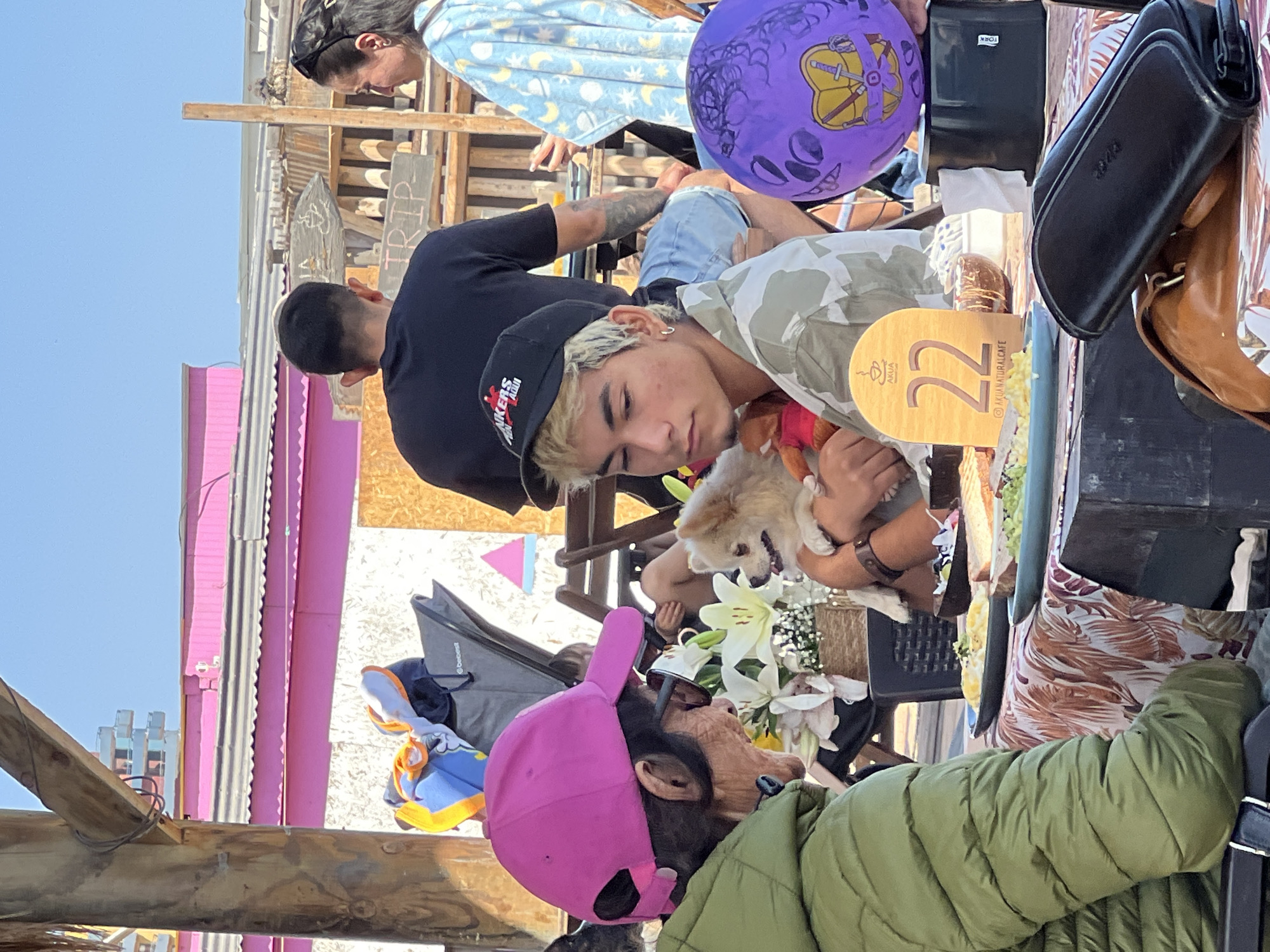 The image depicts an outdoor scene, likely a cafe or restaurant, in Arica, Chile. Four people are visible: one wearing a black t-shirt, another a light blue top with moons, a third wearing a green puffer jacket and a pink cap, and a young man wearing a camouflage shirt and a black cap. The young man is holding a small, fluffy dog. A table displays a number "22" on it, along with an "AKUA natural cafe" logo, indicating the name of the establishment. There is also a purple balloon and a black purse on the table. The background includes elements of a wooden structure, possibly the restaurant's exterior, and glimpses of the city's architecture.