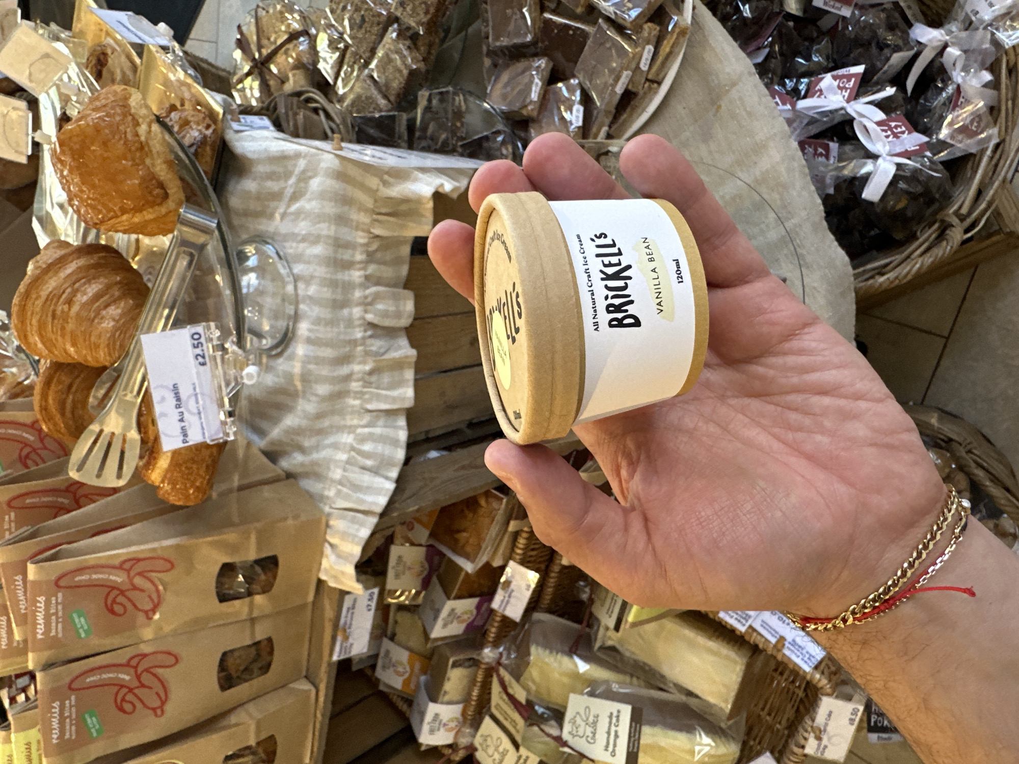 A hand holds a small container of Brickell's Vanilla Bean ice cream (120ml) against a backdrop of various pastries and packaged goods in a store setting. The pastries include croissants, pain au raisin, and other baked items. Many of these items are displayed with price tags (€2.50, €8.50, etc.). The hand wearing a gold bracelet with a red thread is likely that of a customer in a bakery or confectionery shop. The handwritten style labels suggest the products are handmade. The scene suggests a retail environment, possibly a local food market or artisan bakery.