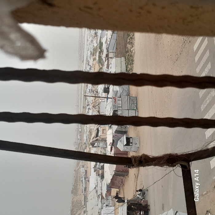 A view through a window or bars shows a refugee camp. Makeshift shelters, tents, and simple structures are scattered across a barren landscape. A road with a pedestrian crossing runs beside the camp. The scene has a hazy, overcast quality, suggesting a bleak and desolate environment. The bars or window frame in the foreground provide a sense of confinement, highlighting the precariousness of the camp's inhabitants.