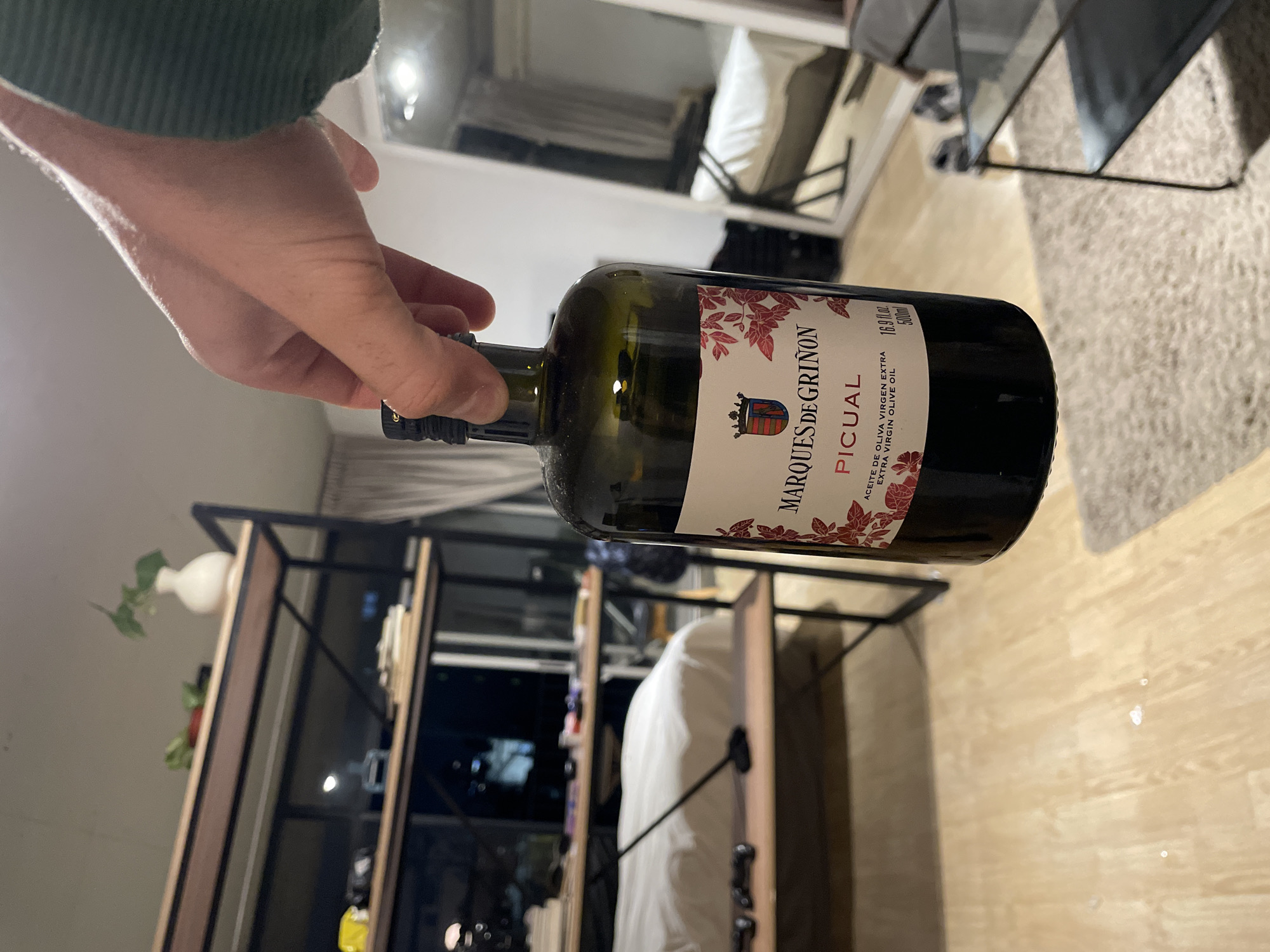 A hand is holding a bottle of Marqués de Griñon Picual extra virgin olive oil. The bottle is dark green glass with a white label featuring a red and brown logo and text. The room contains various furnishings, including a bookshelf-style unit with dark metal framing and light brown wooden shelving, and a chair or stool with a dark metal frame and a light cushion. Other furniture and a window are visible in the background. The setting may be in a private residence within the city of Buenos Aires, Argentina. The presence of natural lighting suggests a daytime shot.