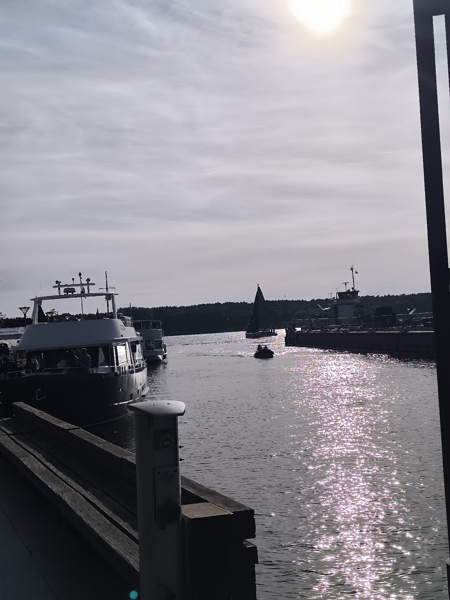 The image depicts a harbor scene in Klaipėda, Lithuania. The foreground shows a wooden pier with a metallic bollard. Moored to the pier is a large, white passenger boat, with several people visible on its deck. In the background, the water is sparkling with sunlight. Further out in the harbor, other boats are visible, including a sailboat with dark sails and a smaller motorboat with occupants. The horizon is lined with a dense forest. The sky is overcast with scattered clouds, and the sun is visible, creating a bright glare. The overall impression is of a calm day in a coastal city.