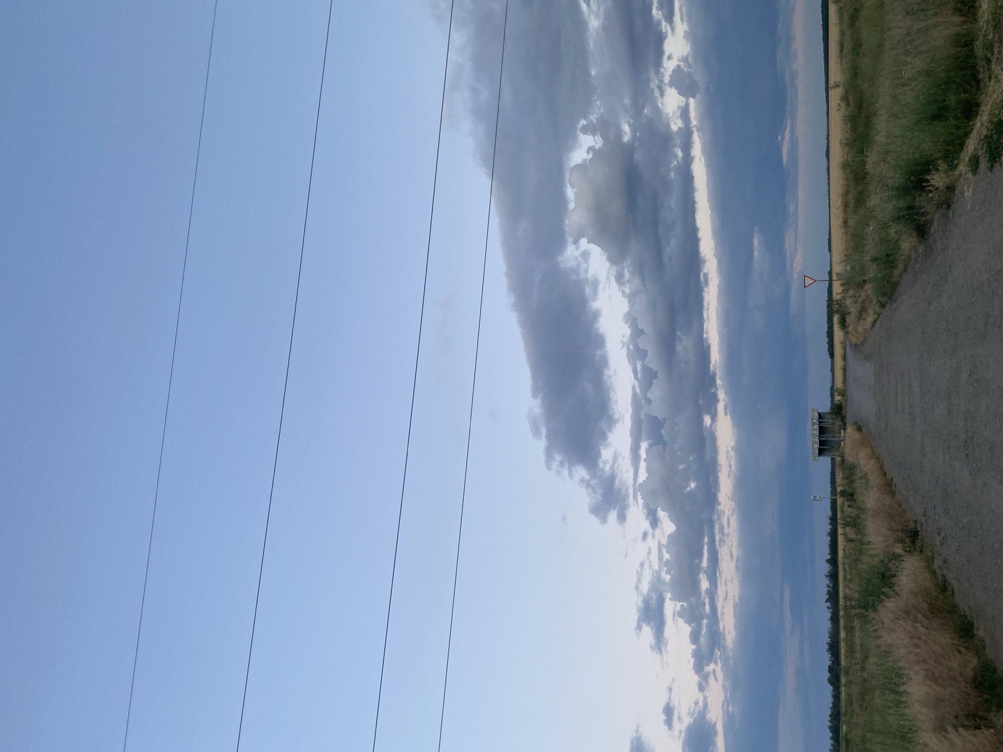 The image depicts a rural landscape under a sky with scattered clouds. In the foreground, several power lines run horizontally across the frame from left to right. The sky is predominantly a pale blue, with darker, elongated clouds stretching across the right side of the image.
The setting appears to be a field with dry, tall grass on the right side, bordering a dirt or gravel path. Further in the distance, a thin line of trees is visible, suggesting a rural or semi-rural environment. A small, triangular red and white sign is planted in the field, likely a warning or directional marker. A simple, open-sided structure, possibly a small shelter or a storage shed, is located near the path and the field.
The time of day appears to be late afternoon or early evening, indicated by the soft lighting and the diffused sunlight through the clouds. The weather seems to be partly cloudy, with a sense of calm and open space. There is no visible text in the image. The overall impression is one of a quiet, natural setting.