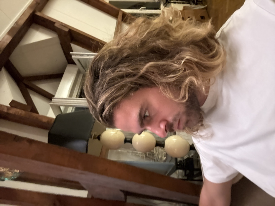 The image portrays a male individual, seen from the chest up in a three-quarters profile, looking left. He possesses shoulder-length, wavy, dirty blonde hair and a short beard, wearing a plain white t-shirt. The setting is an indoor space in Paris, France, specifically an attic or a room with a mansard roof evidenced by the sloped white ceiling and prominent dark brown exposed wooden support beams. Behind the subject, a floor lamp with a black rectangular shade and a stem comprising three stacked light-colored spherical elements is visible. Further back, a white-framed window appears partially open. The environment is dimly lit, suggesting artificial illumination.
