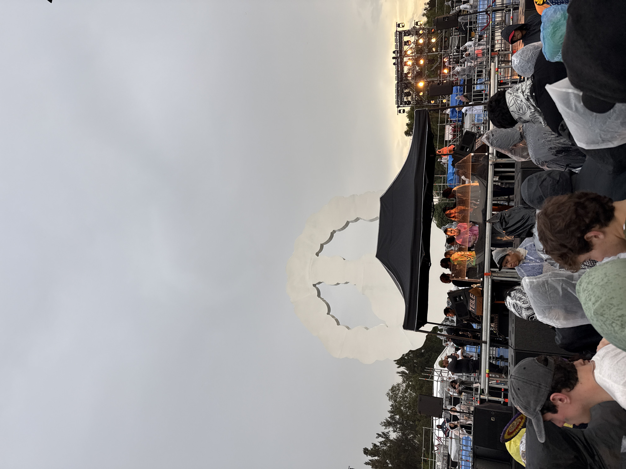 The image depicts an outdoor gathering with a stage and a crowd of people. The sky is overcast with a hint of sunset light peeking through. In the background, a large, white, abstract sculpture resembling a peace sign is visible. A stage setup with lighting equipment and speakers is also present. People are seen standing and watching the stage, many are wearing raincoats, suggesting inclement weather. The setting appears to be an open-air venue. Based on the visual context of a large public event, a stage, and a crowd, this scene could be from a festival or concert event in Mexico City, Mexico.
