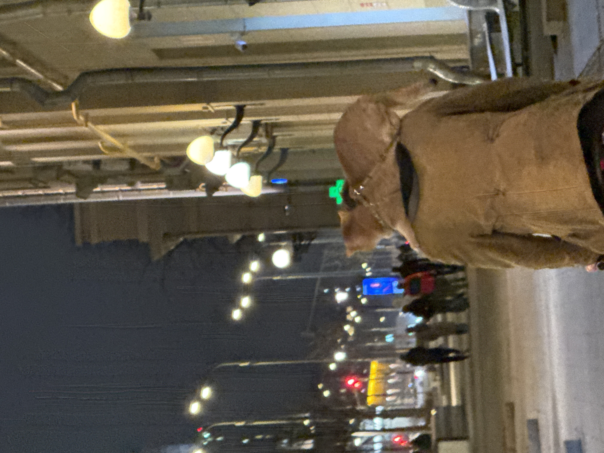 This media file captures a tilted view of an urban street scene at night in Moscow, Russia. A person, viewed from the back and slightly to the side, is prominent in the right foreground, wearing a textured brown or tan coat, possibly suede or fur, with a visible hood. A gold-colored chain or necklace is worn around their neck.
The upper right portion of the image shows an overhead structure, possibly a covered walkway or underpass ceiling, fitted with several bright, warm-toned lights and industrial-style conduits. A light blue horizontal panel on this structure displays faint, unreadable red text.
The background on the left reveals a bustling city street illuminated by numerous artificial lights against a dark night sky. These include blurred streetlights, red traffic lights, and various illuminated signs, notably a bright green square light and a blue rectangular sign. Several other blurred figures, likely pedestrians, are visible further down the street, suggesting an active urban environment. The image conveys the vibrant atmosphere of a city after dark.