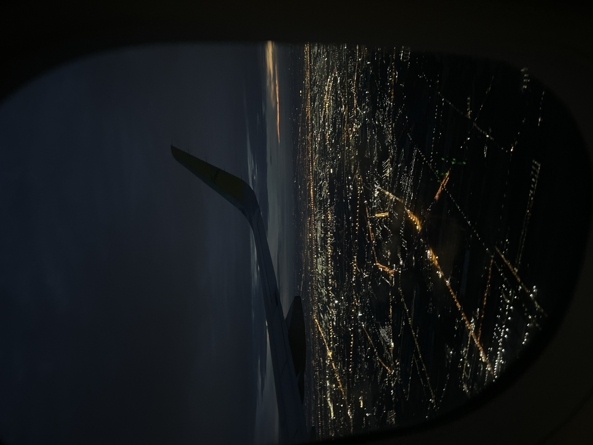 An aerial view of the city of Bangkok, Thailand, is seen through the window of an airplane during nighttime. The city's illumination creates a network of light patterns, and the plane's wing is partially visible in the view.