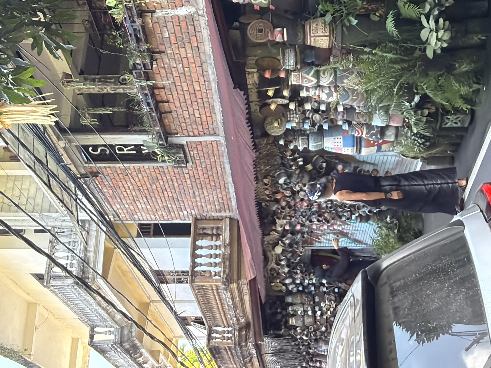 A low-angle, medium shot captures the exterior of a shop in Ubud District, Indonesia, filled with decorative clay pots and other crafts. The shop's facade is adorned with a variety of earthenware, stacked and displayed, creating a dense visual texture. A deep red awning extends over the display area. Above the shop, a building with balconies features elaborate railings and cream-colored walls, contrasting with the red brick of the shop's upper story. Overhead, a tangle of electrical wires crisscrosses the sky.
In the foreground, the rear of a dark-colored SUV is visible, with its rear windshield showing a reflection of what appears to be foliage and the sky. Two individuals are present. One, on the left, wears dark clothing and appears to be interacting with the shop's display. The second individual, on the right, stands with their back to the camera, also dressed in dark clothing, including a long skirt and a sleeveless top, and wears a headscarf. They are looking towards the shop. Lush green plants and ferns are interspersed among the displayed items, adding a touch of nature to the scene. A decorative yellow fringe hangs from a structure above the shop, possibly a cultural ornament. The overall impression is of a vibrant, artisanal marketplace in a tropical urban setting.