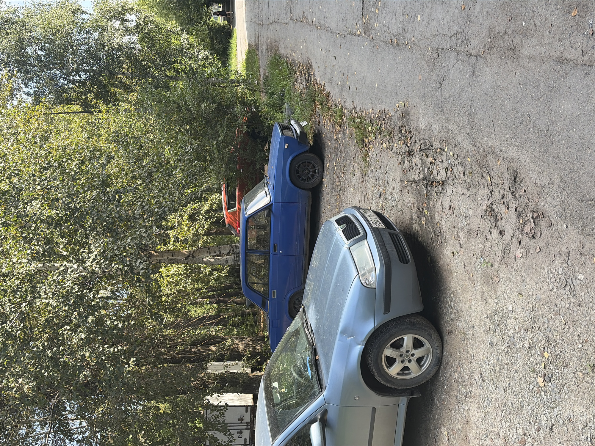 The image captures a street scene in Yekaterinburg, Russia, characterized by parked vehicles and natural surroundings. In the foreground, a light blue sedan, possibly a Skoda Octavia, is parked on a gravel surface. To its right and slightly behind, a blue sedan, likely a Lada, is also parked. A glimpse of a red vehicle is visible behind the blue Lada. The scene is bordered by a line of trees with dense foliage, suggesting a park or a residential area with ample greenery. A paved road runs along the right side of the image, with a person walking away from the camera on the far right. The ground is a mix of gravel and dirt, with fallen leaves scattered around, indicating an autumn or late summer setting. The lighting suggests it is daytime, with sunlight filtering through the trees.