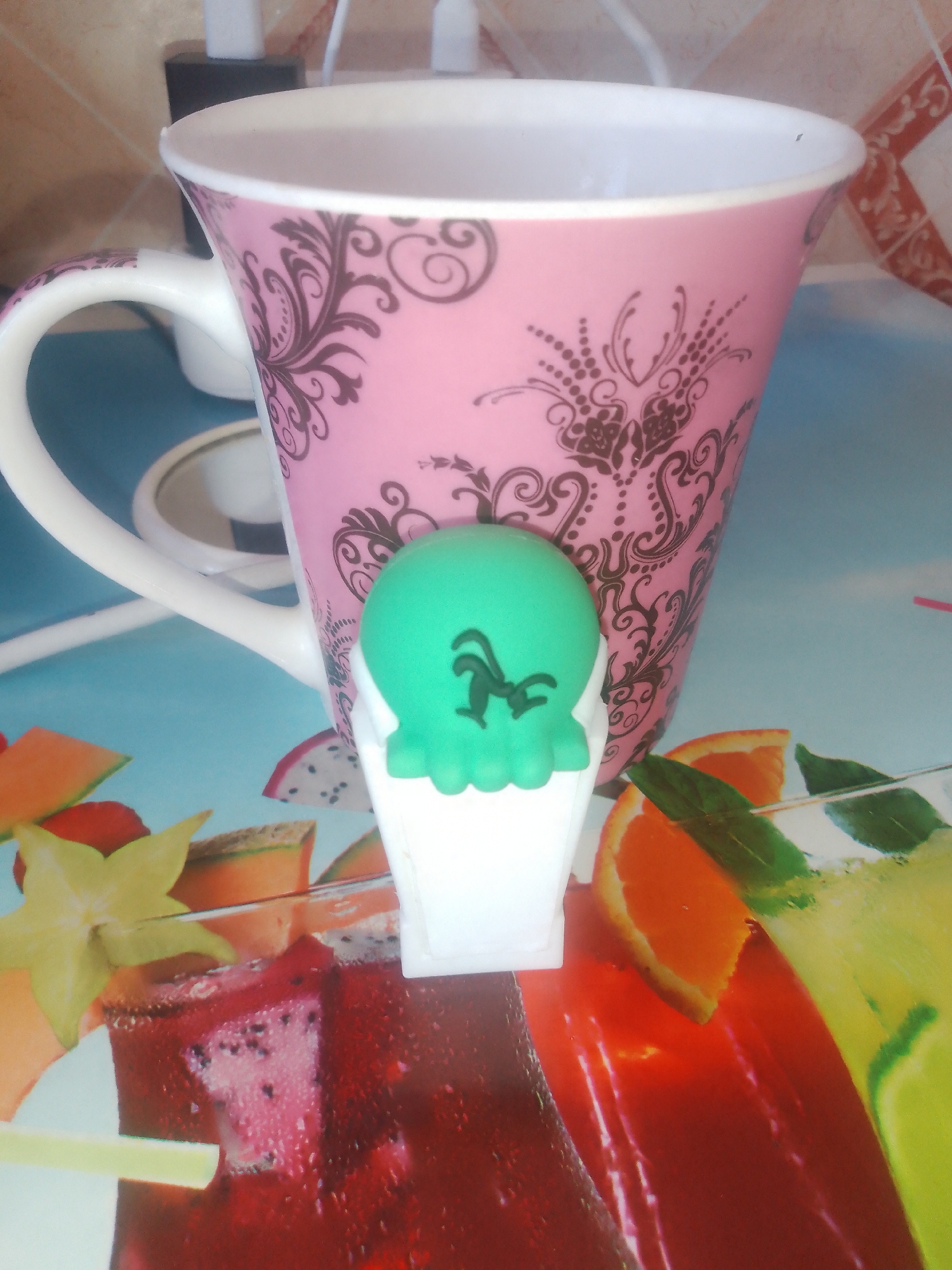 The image shows a pink mug with a brown floral pattern and a white handle. In front of the mug is a green toy that resembles an ice cream cone with a face. The mug is placed on a surface that appears to be a table or placemat with a colorful pattern of fruits and drinks, including slices of orange, lime, and possibly dragon fruit and cantaloupe. There are also images of what look like glasses of juice or cocktails. In the background, a white electrical outlet is visible. The lighting suggests an indoor setting, possibly a kitchen or dining area. There is no information or visual cues to indicate this scene is located in Chernivtsi, Ukraine.