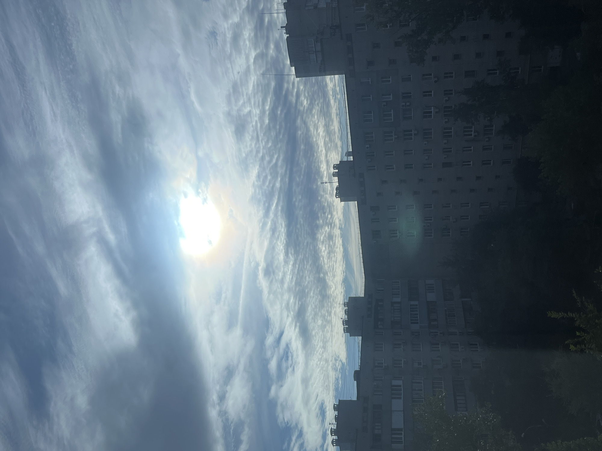 The image depicts a view of apartment buildings in Volgograd, Russia, under a cloudy sky with the sun partially obscured. The buildings appear to be typical Soviet-era residential blocks, characterized by their uniform grey facades and numerous windows. Some of the buildings have chimneys on their roofs. Trees and foliage are visible in the foreground and between the buildings, suggesting an urban or suburban setting. The lighting indicates it is daytime, possibly late morning or early afternoon, with the sun's rays filtering through the clouds, creating a diffused light. There are no people or visible actions in the scene, it is a static environmental view.