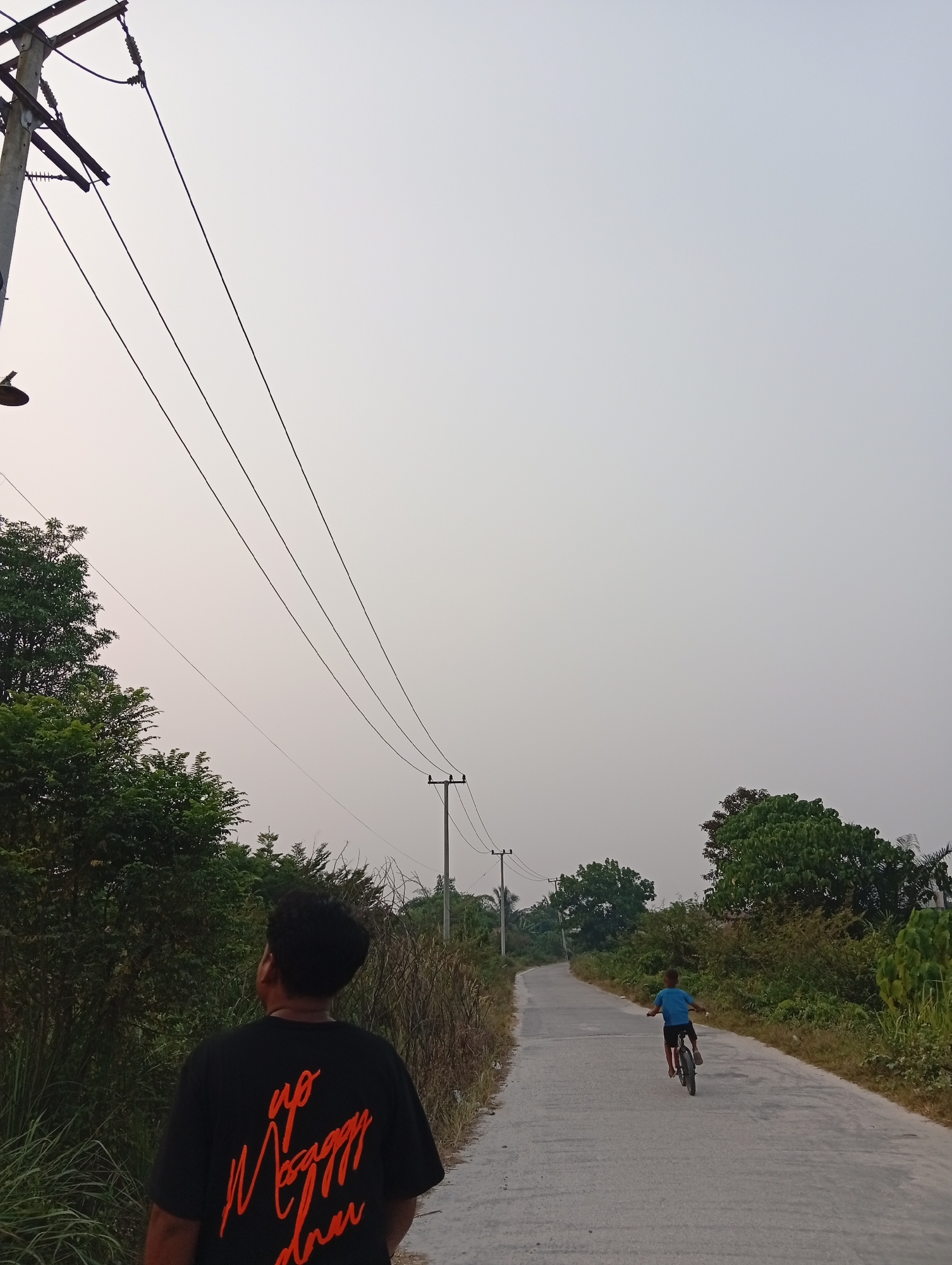 In Kualu, Indonesia, on a cloudy day, a paved road winds through a rural landscape bordered by lush green vegetation and utility poles with suspended wires. In the foreground, a young person wearing a black t-shirt with orange lettering faces away from the camera, looking up the road. Further down the road, a child cycles away from the viewer on a bicycle. The scene captures a moment of everyday life in the Indonesian countryside.