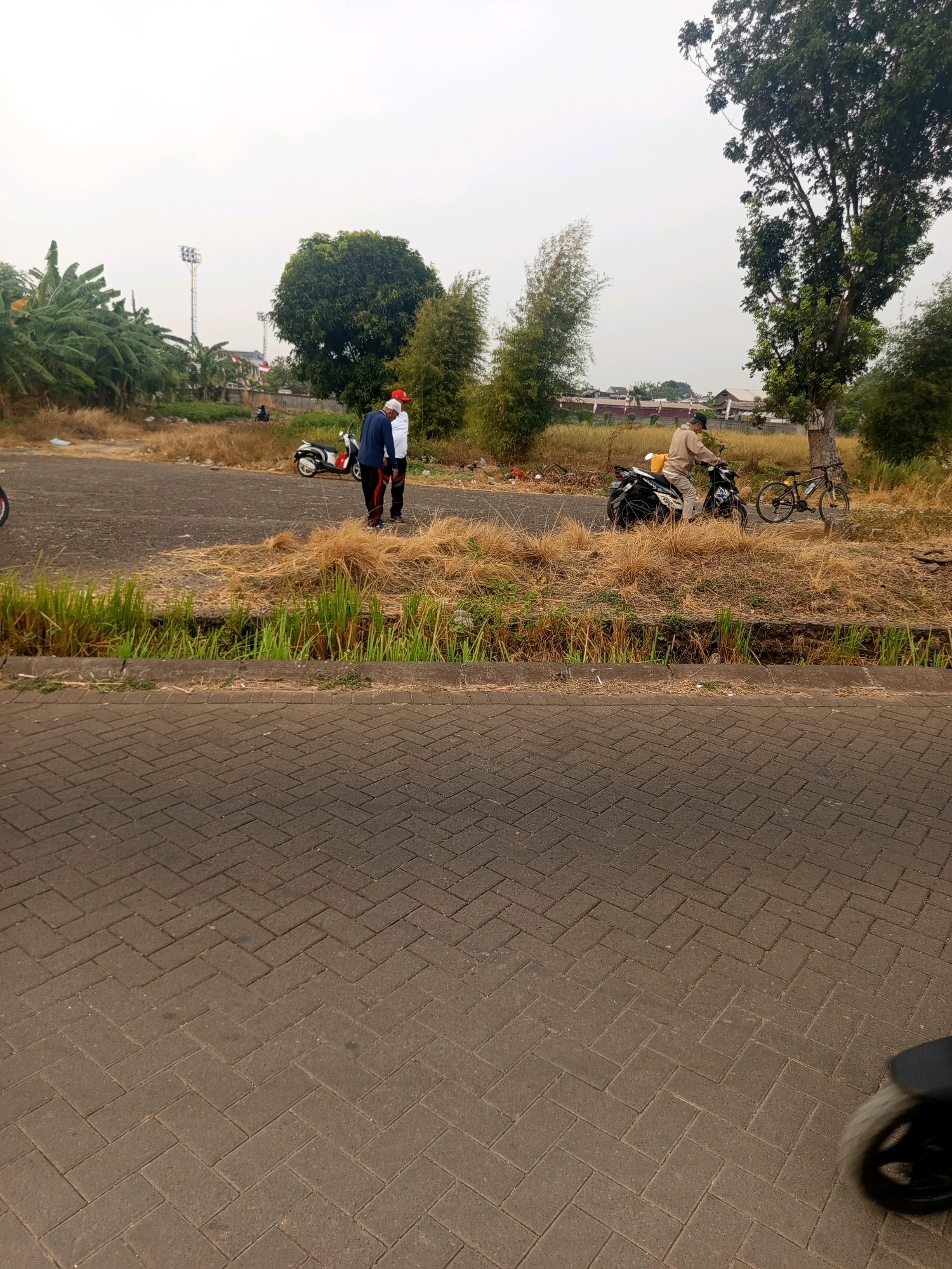 The image depicts an outdoor scene with two men standing near a white scooter and a man on a black motorcycle with a yellow bag on the back. A bicycle is parked behind the motorcycle. The ground in the foreground is paved with herringbone patterned bricks. In the background, there is dry grass, some green vegetation, and a large tree. Two tall stadium lights are visible in the distance. The sky is overcast. The location is a street or parking area in West Jakarta, Indonesia, with a residential or undeveloped area beyond the paved surface.