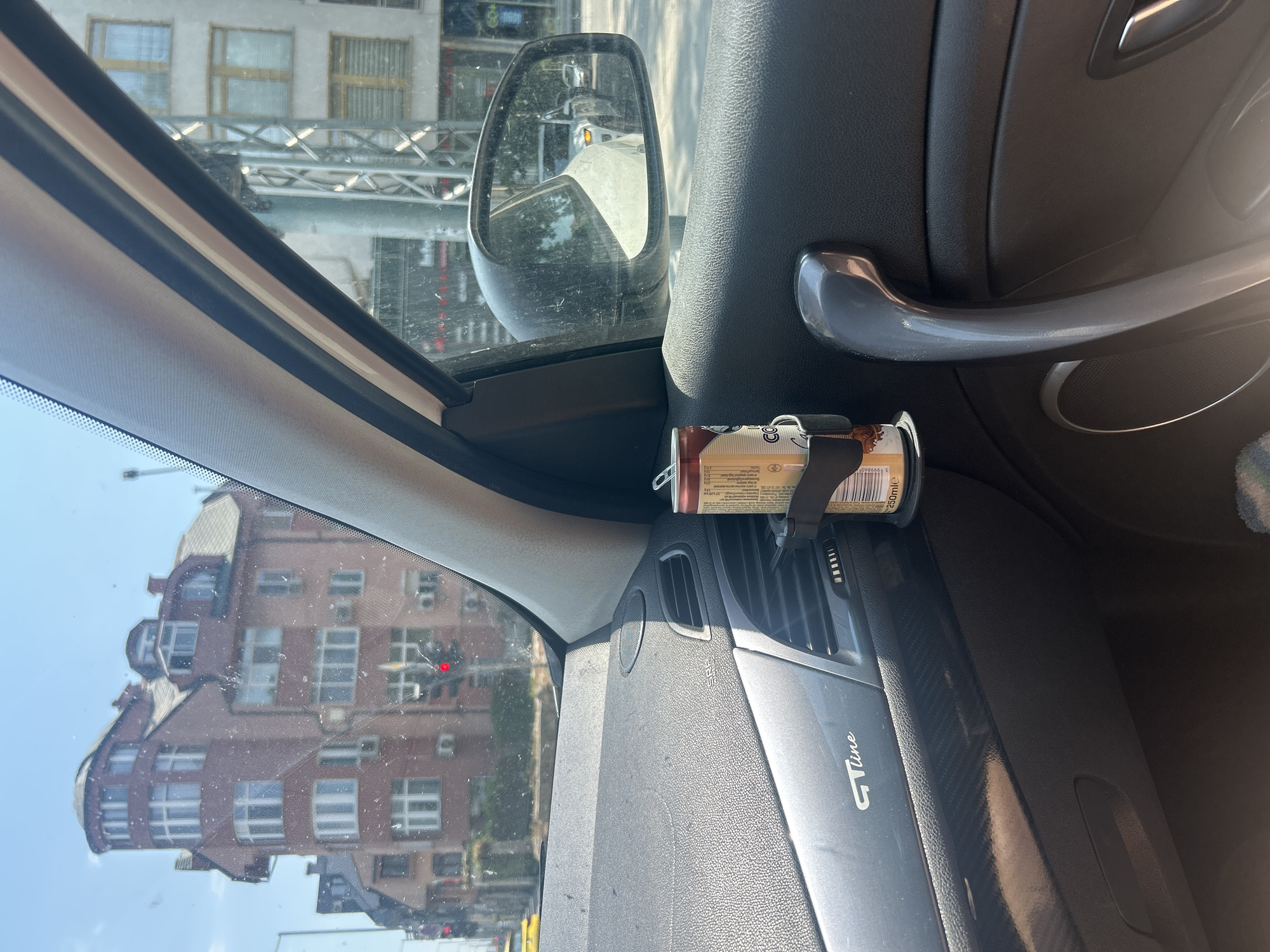 The image captures a view from inside a car in Plovdiv, Bulgaria, looking out the passenger-side window. The focus is on the interior of the car, with a can of what appears to be coffee in a cup holder attached to the air vent. The dashboard is visible on the left, showing the "GT Line" emblem. Outside the car, a red brick building with multiple windows and a distinctive turret dominates the scene, suggesting an urban setting. Traffic lights are visible, one showing red. The side mirror reflects other cars and buildings. The sky is clear and blue.