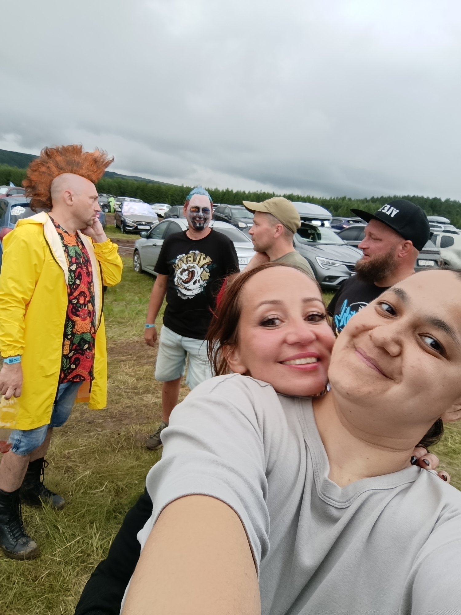 In Lagunovo, Russia, a group of people are gathered outdoors, likely at a festival or event. The foreground is dominated by two women posing for a selfie; one has her arm around the other, both smiling. In the midground, a man with a distinctive orange mohawk and a bright yellow raincoat stands to the left, talking on his phone. To his right, a man with face paint resembling a skull and blue hair wears a black t-shirt and shorts. Further back, a man in a beige baseball cap and another in a black baseball cap with "AJW" printed on it are visible, with several cars parked behind them. The environment appears to be a grassy field with a cloudy sky overhead.
