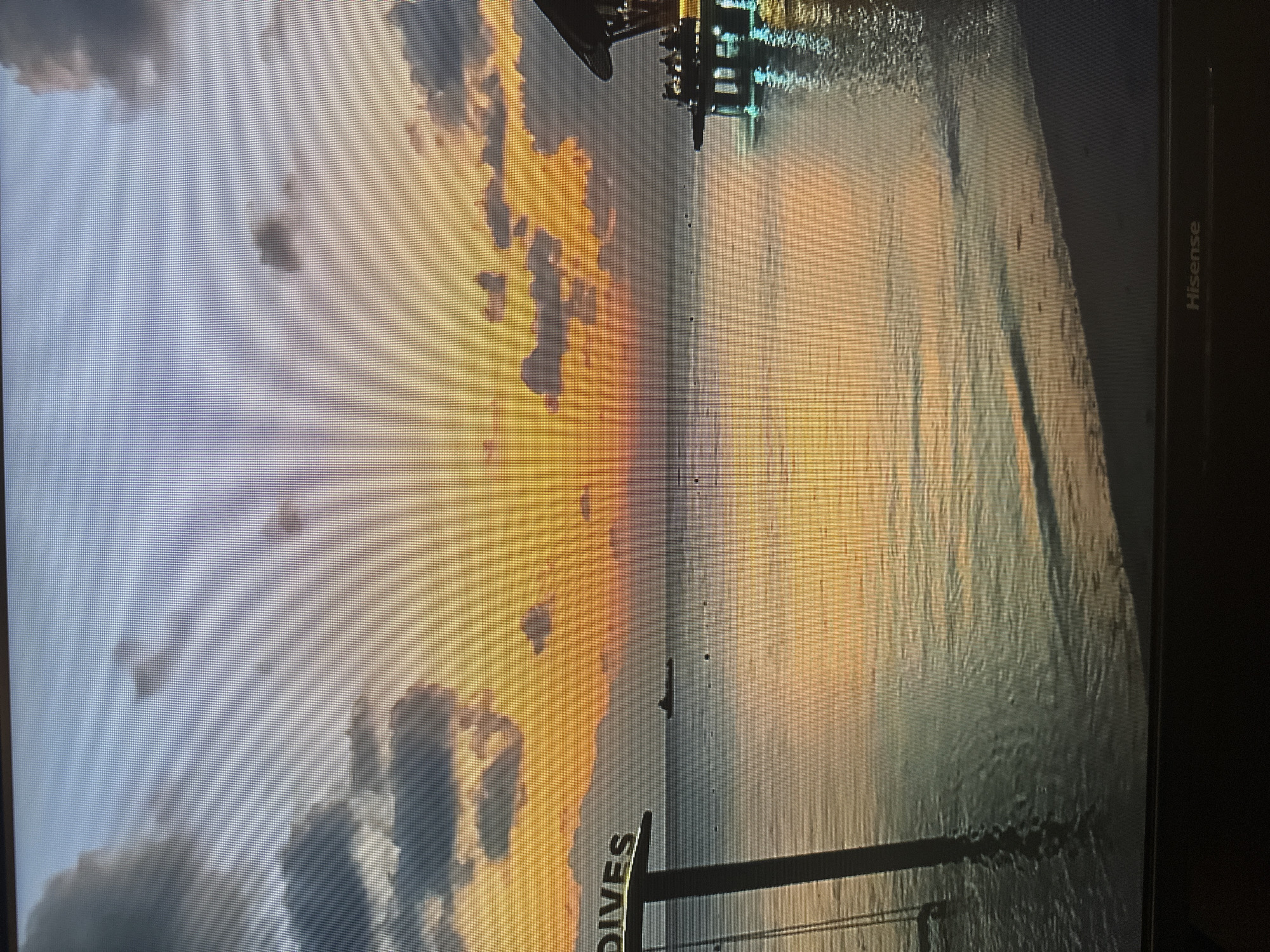 The image shows a serene sunset over a body of water, likely the ocean. The sky is painted with vibrant hues of orange, yellow, and purple, with scattered clouds catching the last rays of the sun. The calm water reflects the colors of the sky, creating a tranquil and picturesque scene. In the foreground, a wooden post with the word "DIVES" is partially submerged in the water, suggesting a diving area or a resort. To the right, a pier with people enjoying the view extends into the water, illuminated by soft lights. A boat is visible in the distance, adding a touch of life to the vast expanse of water. The overall atmosphere is one of peace and relaxation, a perfect ending to a day in a tropical paradise. The location is not in Kyiv, Ukraine, but rather a tropical beach.