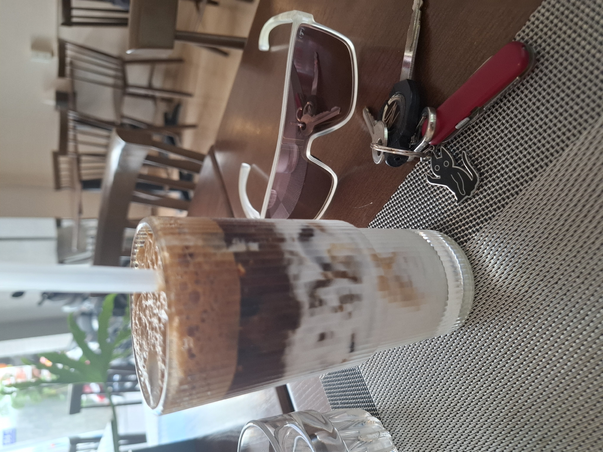 The image shows a table in an outdoor seating area, likely a cafe or restaurant, in Nha Trang, Vietnam. The time of day appears to be daytime, possibly afternoon, given the lighting conditions. The table is set with a tall glass containing a layered drink, likely a dalgona coffee or similar iced beverage, with a white straw.
On the table, next to the drink, are a pair of white-rimmed sunglasses. Also present are a set of keys attached to a keyring, which includes a small red multi-tool (possibly a Swiss Army knife) and a black charm shaped like a cat. The table is covered with a textured placemat. In the background, wooden chairs and tables are visible, suggesting a casual dining or relaxation environment. The overall mood is relaxed and casual. There is no visible text in the image.