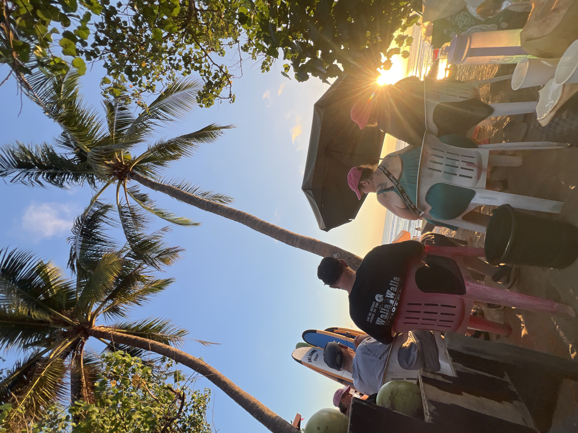 The image depicts a beach scene in Kuta, Indonesia, during sunset. Three individuals are seated in chairs facing the ocean. One person, on the left, wears a pink cap and a green dress. Another, in the center, is seated and partially visible from behind, wearing a black t-shirt with white lettering. The third person, on the right, is also seated, partially obscured by an umbrella. In the background, surfboards are propped up, and palm trees frame the scene. The sun is setting over the ocean, casting a warm glow and lens flare across the image. In the foreground, there are coconuts and cups on a table.