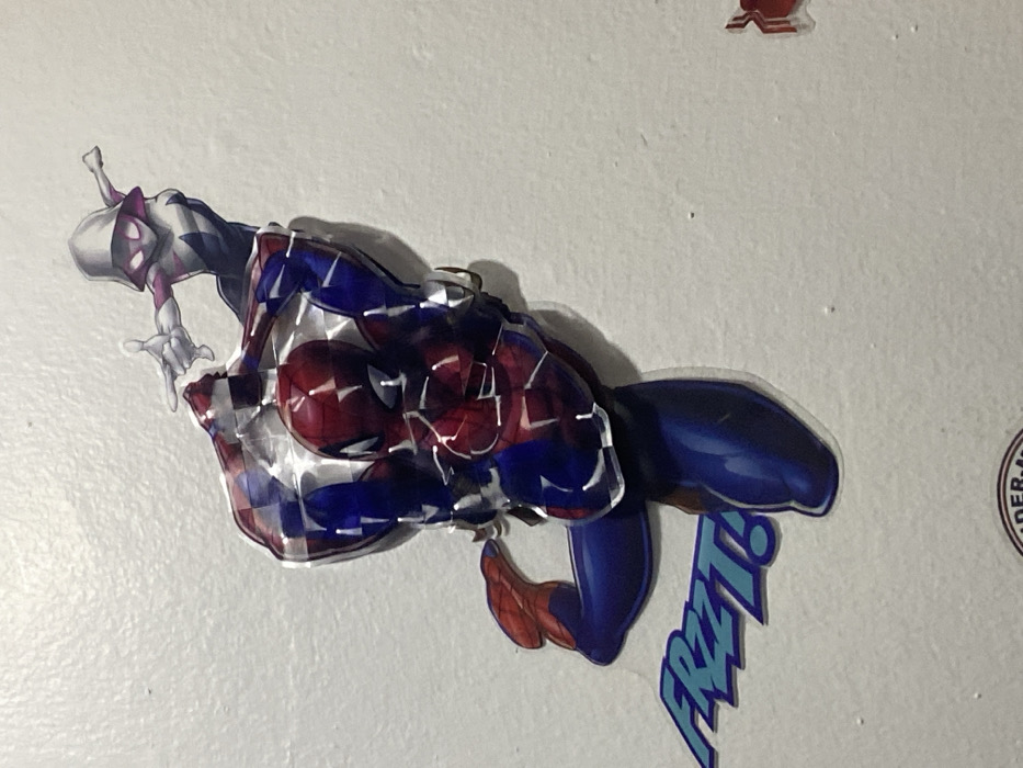 The scene depicts an interior wall located in Tema, Ghana, adorned with several graphic decals. A central, multi-layered decal portrays Spider-Man in his classic red and blue costume, positioned in a dynamic, ascending or grappling pose. The torso of the Spider-Man figure exhibits a faceted, reflective surface, creating a three-dimensional effect. Above and to the upper right of the Spider-Man decal, a flat decal of Spider-Gwen is affixed, rendered in a white, black, and pink hooded suit, with her right arm extended forward. Directly below the Spider-Man decal, a blue text decal reading "FREEZ!" is present in a stylized font. The lower edge of the image partially reveals a circular logo or emblem. The wall surface is uniformly light-colored, indicative of white or off-white paint, and displays minor textural irregularities along with a few small, dark specks.