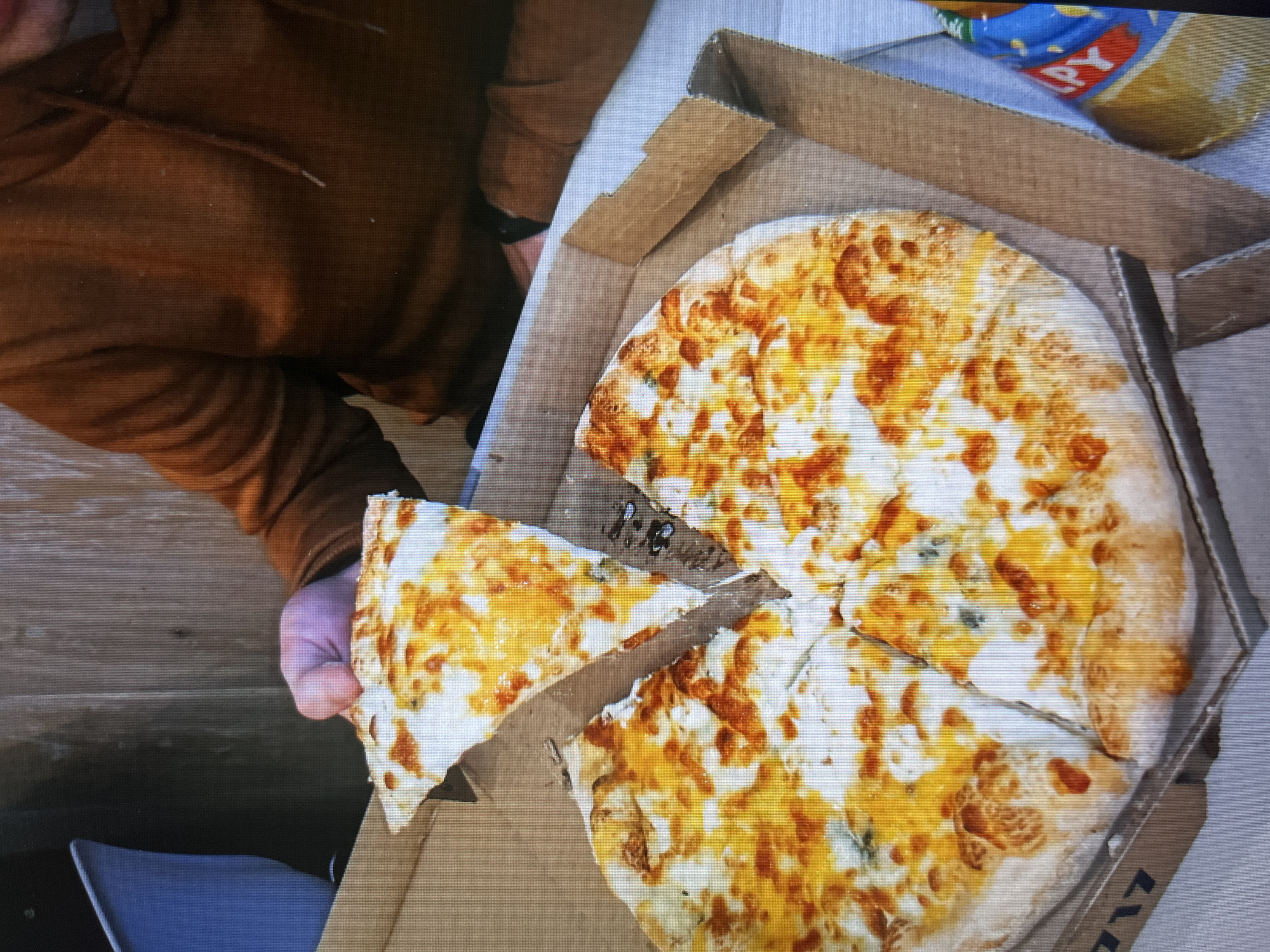 A person wearing a brown hoodie is holding a slice of pizza from a cardboard pizza box containing the rest of the pizza. The pizza is mostly white with orange and yellow toppings. A bag of chips is visible in the upper right corner of the image. The location and city are not discernible from the image.