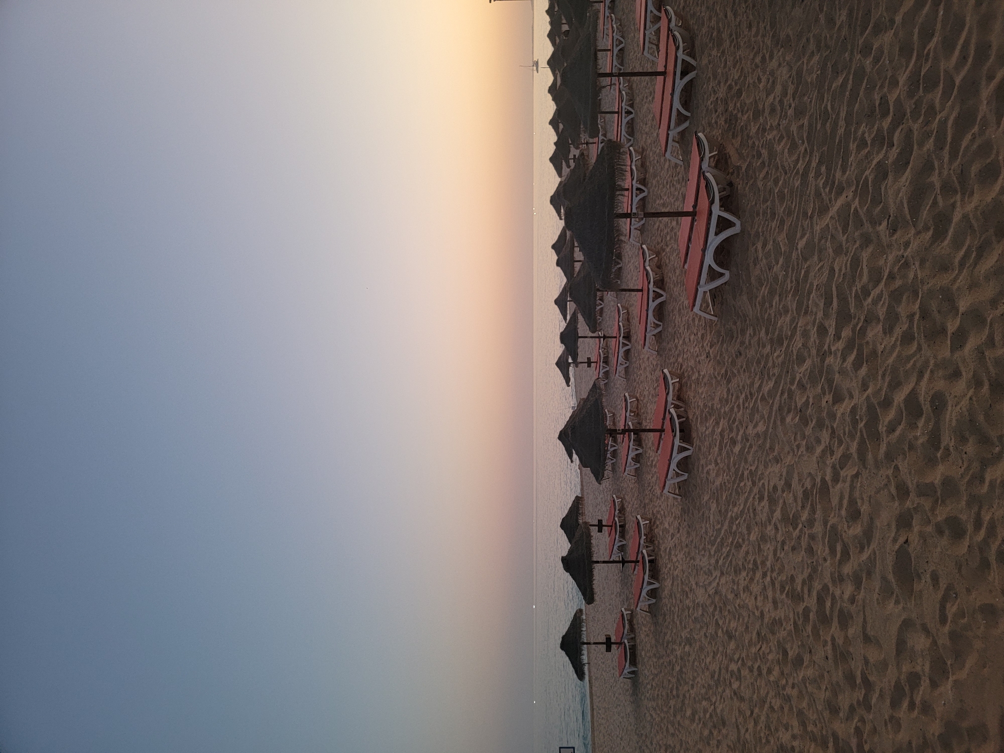 The image shows a beach in Quarteira, Portugal, at dusk. The sky is a gradient of pale blue to soft orange, with the ocean meeting the horizon. The sand is a light brown color and is mostly empty, with a few rows of empty lounge chairs and thatched umbrellas neatly arranged. The chairs are white with orange cushions. In the distance, a small sailboat is visible on the water. The overall atmosphere is peaceful and serene.