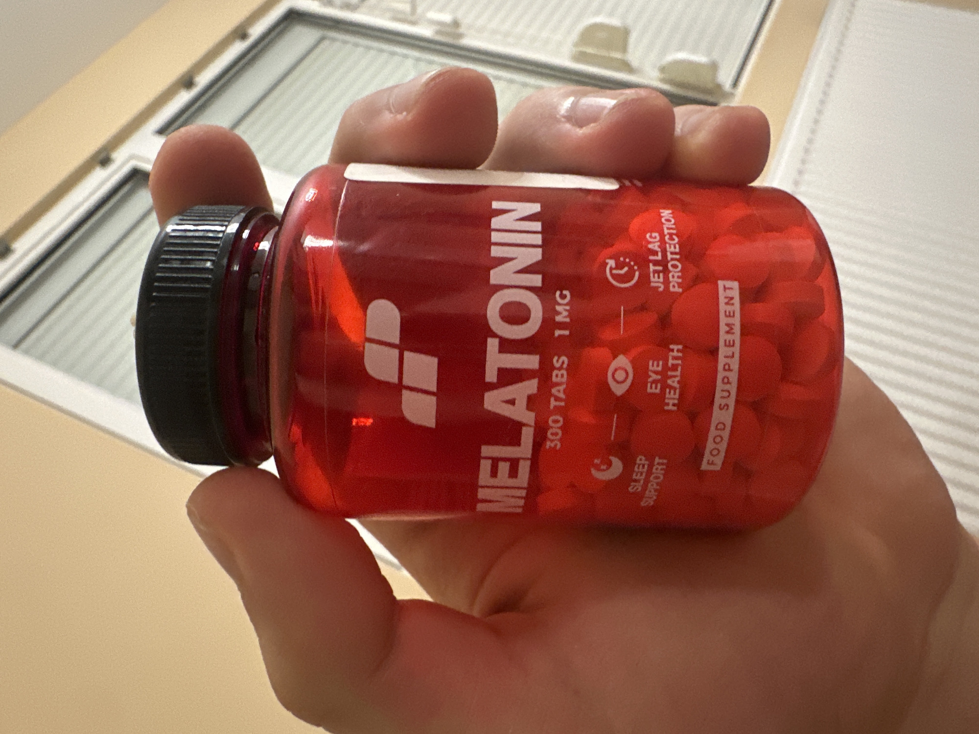 A person's hand holds a red plastic bottle of Melatonin pills, captured indoors in Blagoevgrad, Bulgaria. The bottle is positioned horizontally, grasped by the fingers and thumb of a light-skinned hand, dominating the foreground. Inside the transparent red container, numerous small, round, red pills are visible. The bottle is sealed with a black, ribbed screw-on cap.
The bottle's label features white text and icons. The main product name, "MELATONIN," is displayed prominently. Additional text indicates "300 TABS 1 MG" and labels it as a "FOOD SUPPLEMENT." Product benefits are listed alongside corresponding white icons: a circular arrow for "JET LAG PROTECTION," an eye for "EYE HEALTH," and a crescent moon-like symbol for "SLEEP SUPPORT." A stylized white logo, resembling an initial or abstract design, is also visible on the label.
The background is out of focus, revealing an indoor setting with parts of a window. White horizontal blinds or louvers are visible through the window, suggesting natural light entering the room. A light-colored, possibly beige, wall or ceiling is also in the background. The image focuses on the product and the act of holding it, with no specific activity or emotions discernible.
