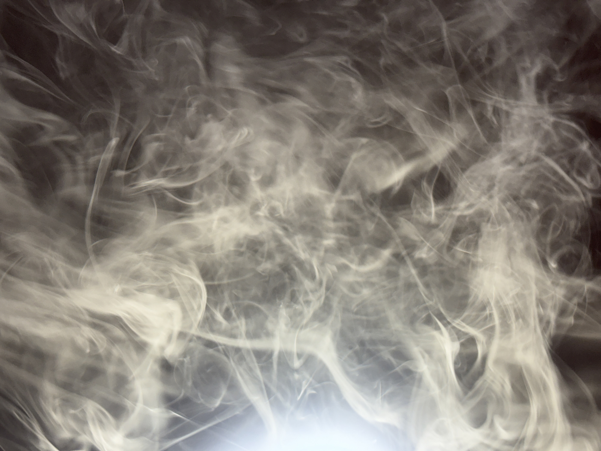 The image shows a swirling pattern of light gray smoke against a dark background. The source of the smoke appears to be from the bottom of the image, where a bright light source illuminates the smoke. There are no people or identifiable objects within the frame. The location of Прикамский, Russia is not relevant to the visible content of the image.