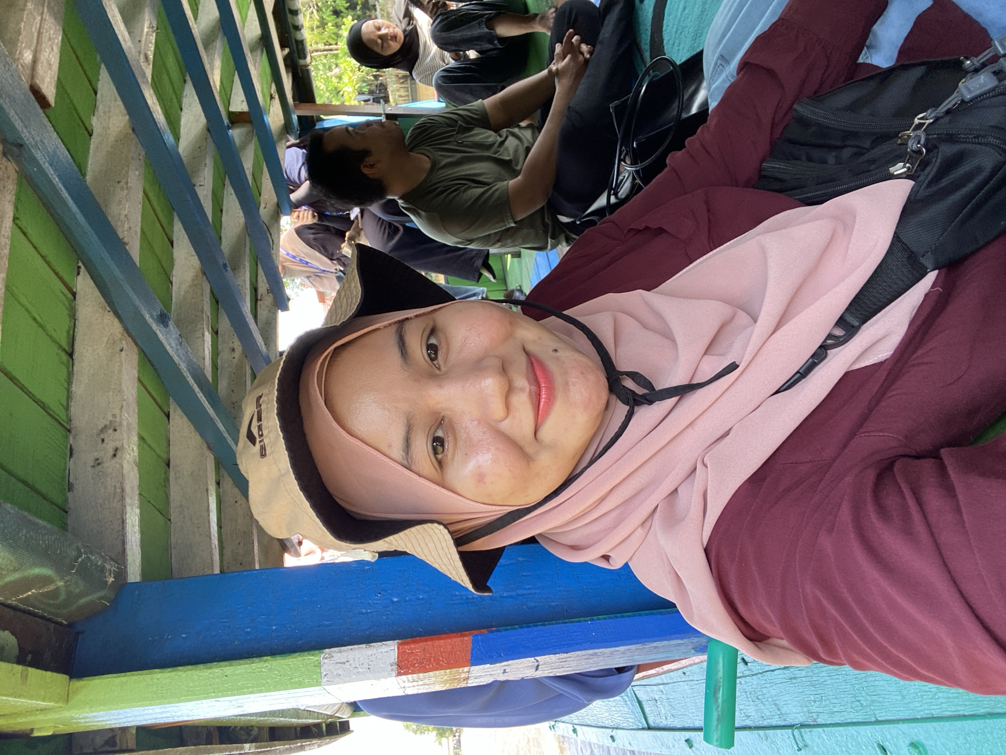 The image shows a group of people on a boat. The foreground is dominated by a woman wearing a pink hijab and a maroon shirt. She is smiling and looking at the camera, and appears to be taking a selfie. Behind her, a man in a green t-shirt is seated, and to his right, a woman in a black hijab is looking at her phone. The boat's interior is visible, with painted wooden beams and planks forming the roof and support structures. The colors are vibrant, with green and blue paint on the ceiling and structural elements. The overall impression is of a casual outing on the water. The location is identified as Banjarmasin, Indonesia, a city known for its extensive river system and floating markets, suggesting this boat trip is likely on one of its many waterways.