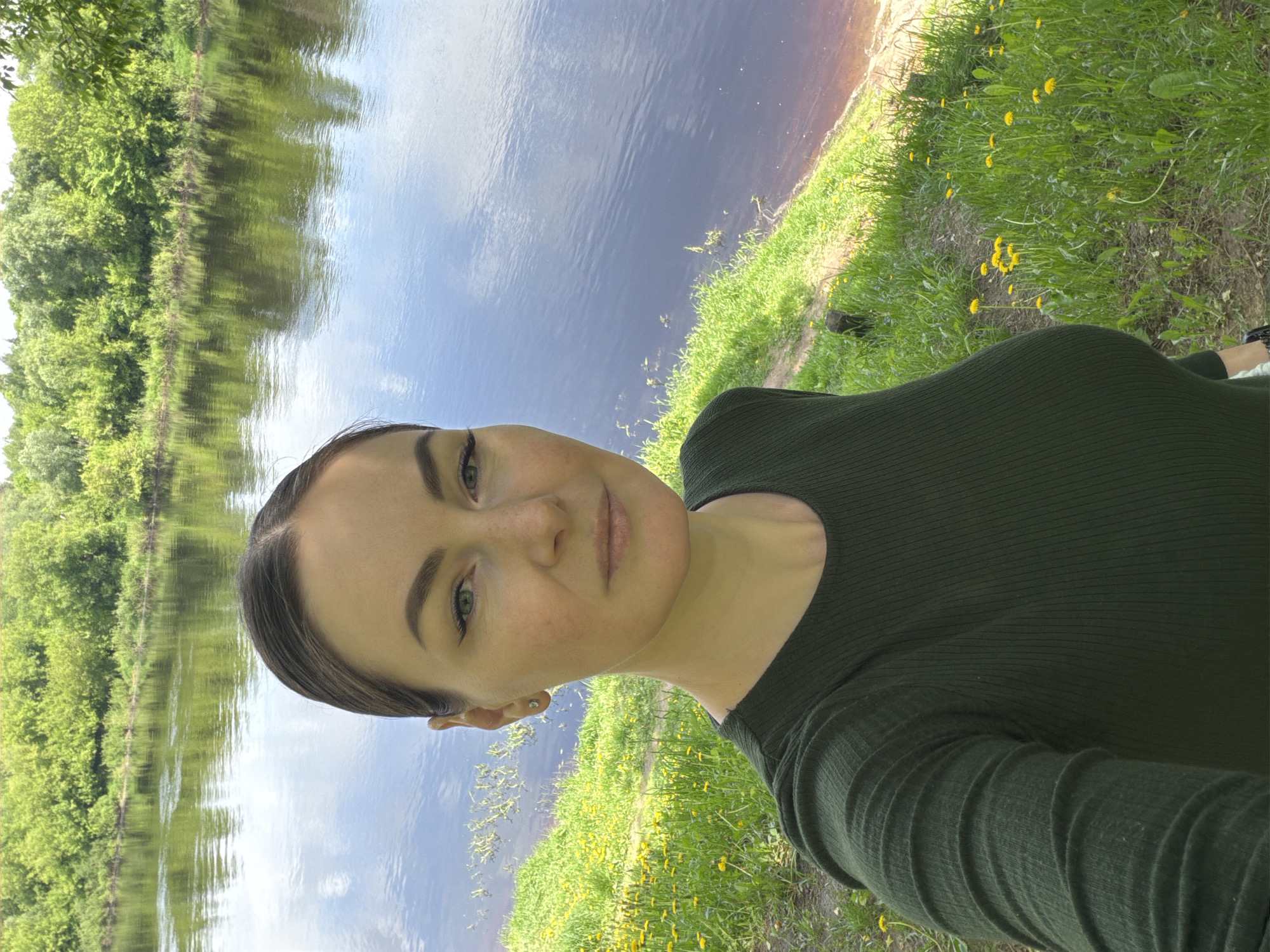The image is a selfie of a woman with fair skin and light brown hair pulled back. She's wearing a dark green long-sleeved shirt. She is standing near a body of water, potentially a river or lake, which reflects the sky and surrounding trees. The foreground consists of green grass interspersed with yellow wildflowers. The location is in the city of Unknown, Unknown.
