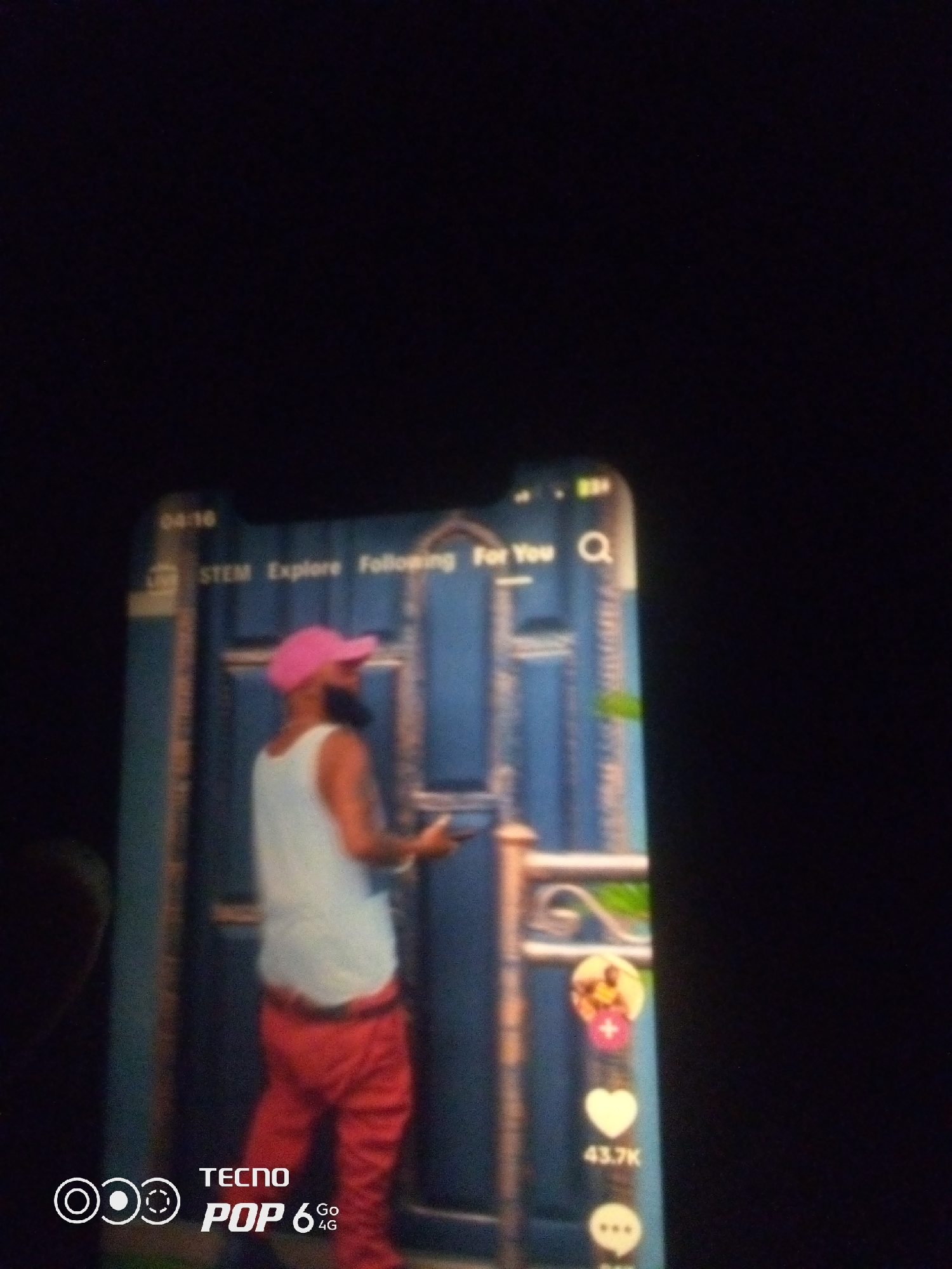 The image shows a vertical screen display, likely a smartphone, showcasing a video. The video features a person wearing a pink baseball cap, a white tank top, and red pants, standing in front of a blue door. The screen displays interface elements typical of a social media application, including text labels like "STEM," "Explore," "Following," and "For You," along with icons for search, profile pictures, likes, and comments. The time "04:16" is visible in the top left corner. The bottom left of the screen displays the "TECNO POP 6 GO 4G" logo. The overall scene is dimly lit, with the bright screen being the primary light source. There is no discernible information within the image to indicate its specific location within the city of Howayi, Nigeria.