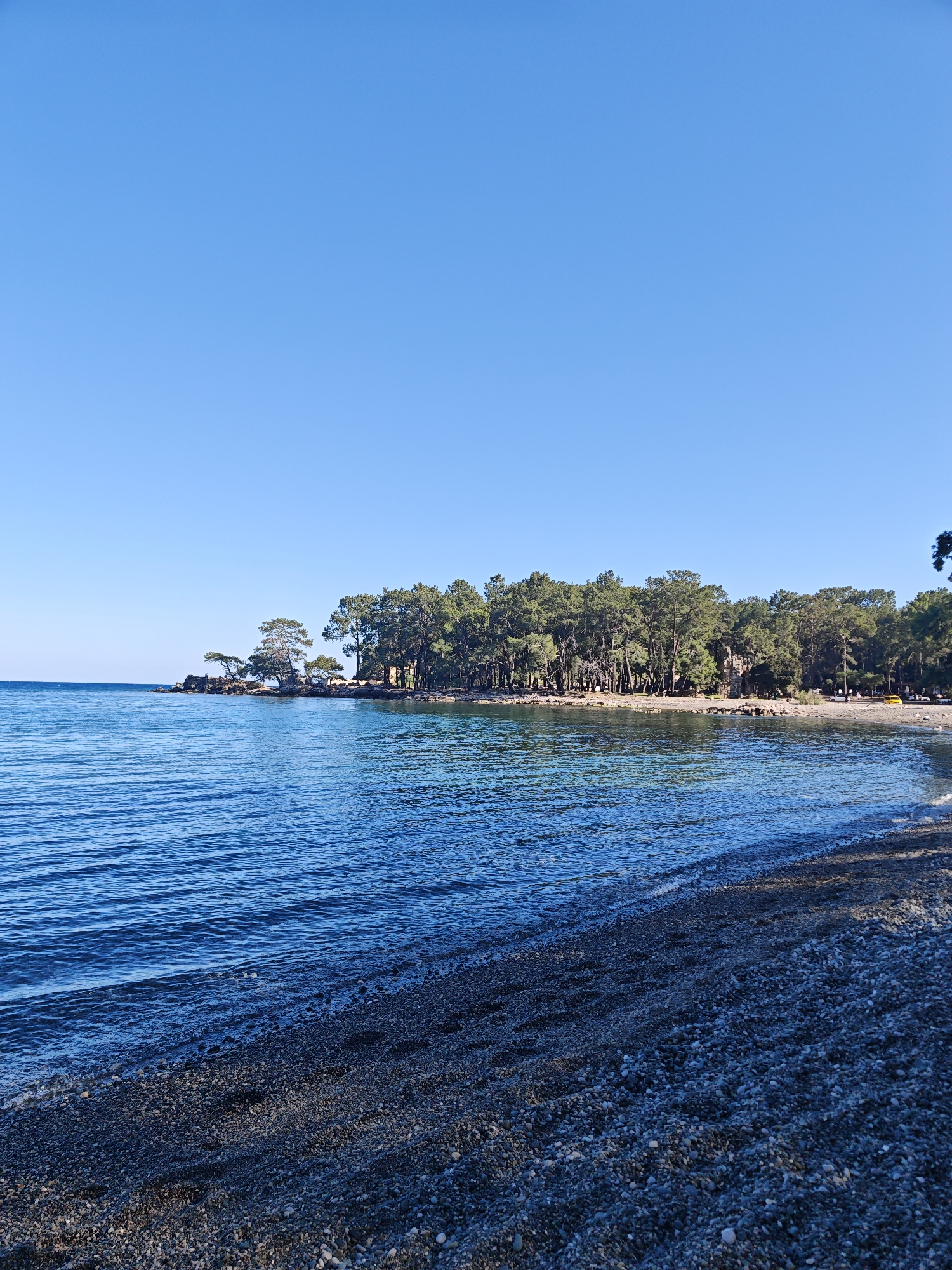 The image presents a serene coastal landscape, likely a tranquil bay, under a vast and clear blue sky, suggesting a bright and sunny day, possibly morning or early afternoon. The setting is Kemer, Turkey.
The scene is dominated by the calm, clear blue-green waters of the sea in the foreground and midground, exhibiting gentle ripples and subtle reflections. On the left, the water extends to the horizon. The immediate foreground features a dark pebble and gravel beach, where the water softly meets the shore. The beach curves along the bay, extending into the distance on the right side of the frame.
The shoreline is densely forested with numerous evergreen trees, likely pines or similar conifers, growing right up to the water's edge. On the left side of the bay, a few prominent trees stand on rocky outcrops that extend slightly into the sea. Further along the tree-lined coast to the right, a light-colored structure, possibly an ancient ruin or a building, is visible nestled within the foliage. Nearby on the beach, faint hints of yellow, perhaps from a vehicle or beach equipment, can be discerned.
The overall impression is one of natural beauty and peacefulness, characteristic of a Mediterranean coastal area where forests meet the sea. There are no prominent human figures or activities visible, enhancing the sense of a pristine and untouched natural environment. No visible text is present in the image.