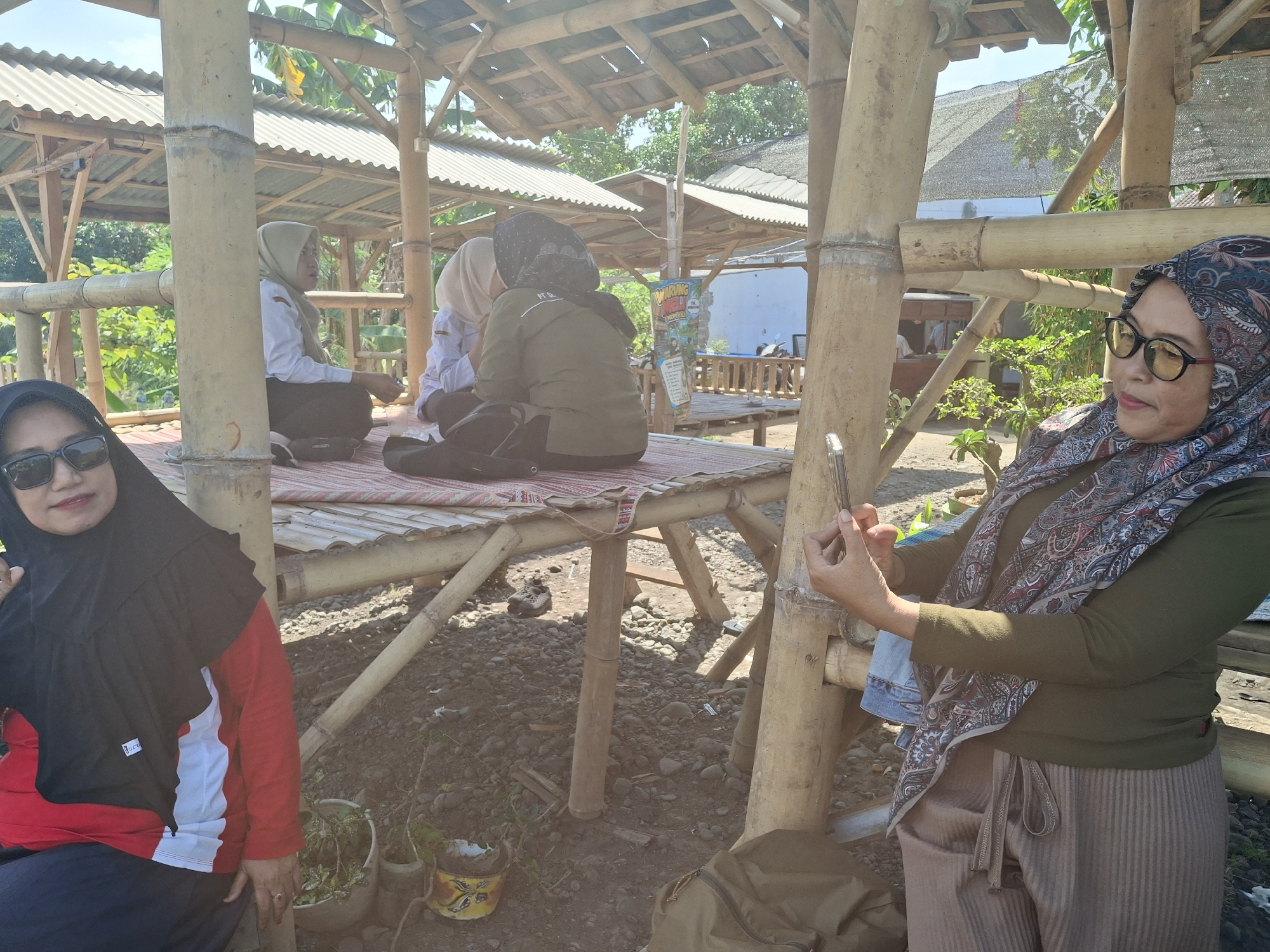 The image shows an outdoor gathering under a bamboo structure with a corrugated metal roof in Lumajang, Indonesia. Four women are present. In the foreground on the left, a woman wearing a black hijab, sunglasses, a red shirt with white and black accents, and dark pants is seated. To her right and slightly further back, three women are seated on a raised platform. One woman in a light-colored hijab and white shirt faces away from the camera. Another woman, wearing a darker green long-sleeved shirt and headscarf, is also seated and facing away. The third woman on the platform, in the foreground on the right, wears a patterned hijab, glasses, a green long-sleeved shirt, and brown striped pants. She is holding a mobile phone in her hands and appears to be using it. The ground is covered with small stones and some potted plants are visible. In the background, there are more bamboo structures and greenery. A sign that reads "WARUNG INDAH" is visible on the platform.