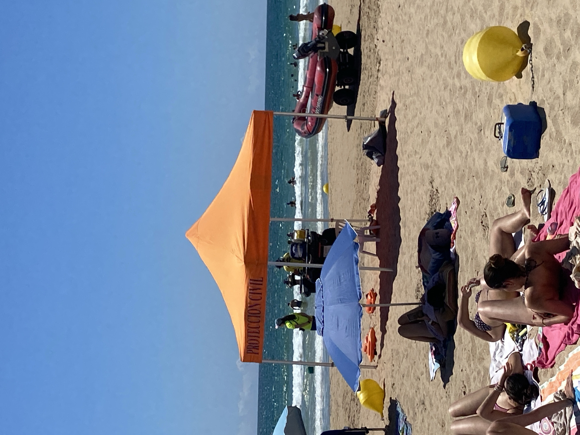 In Conil de la Frontera, Spain, the image depicts a sunny day at the beach with clear blue skies. In the foreground, several individuals are sunbathing on towels and blankets on the sand. Some are lying down, while one person is sitting up and looking at their phone. Further back, a blue beach umbrella provides shade, with two orange life preservers and a white chair underneath.
Behind the umbrella, an orange canopy tent with "PROTECCION CIVIL" written on it stands, indicating a civil protection station. Two individuals in bright yellow vests are visible near a vehicle, and a red inflatable boat with an outboard motor is parked on a wheeled trailer on the sand. In the background, the sea is active with moderate waves, and several people are in the water. Two yellow buoys are also visible in the sea and on the sand. A blue cooler is present on the sand near a yellow cylindrical float. The overall scene is of a typical summer day at a Spanish beach, with recreational activities and safety services present.