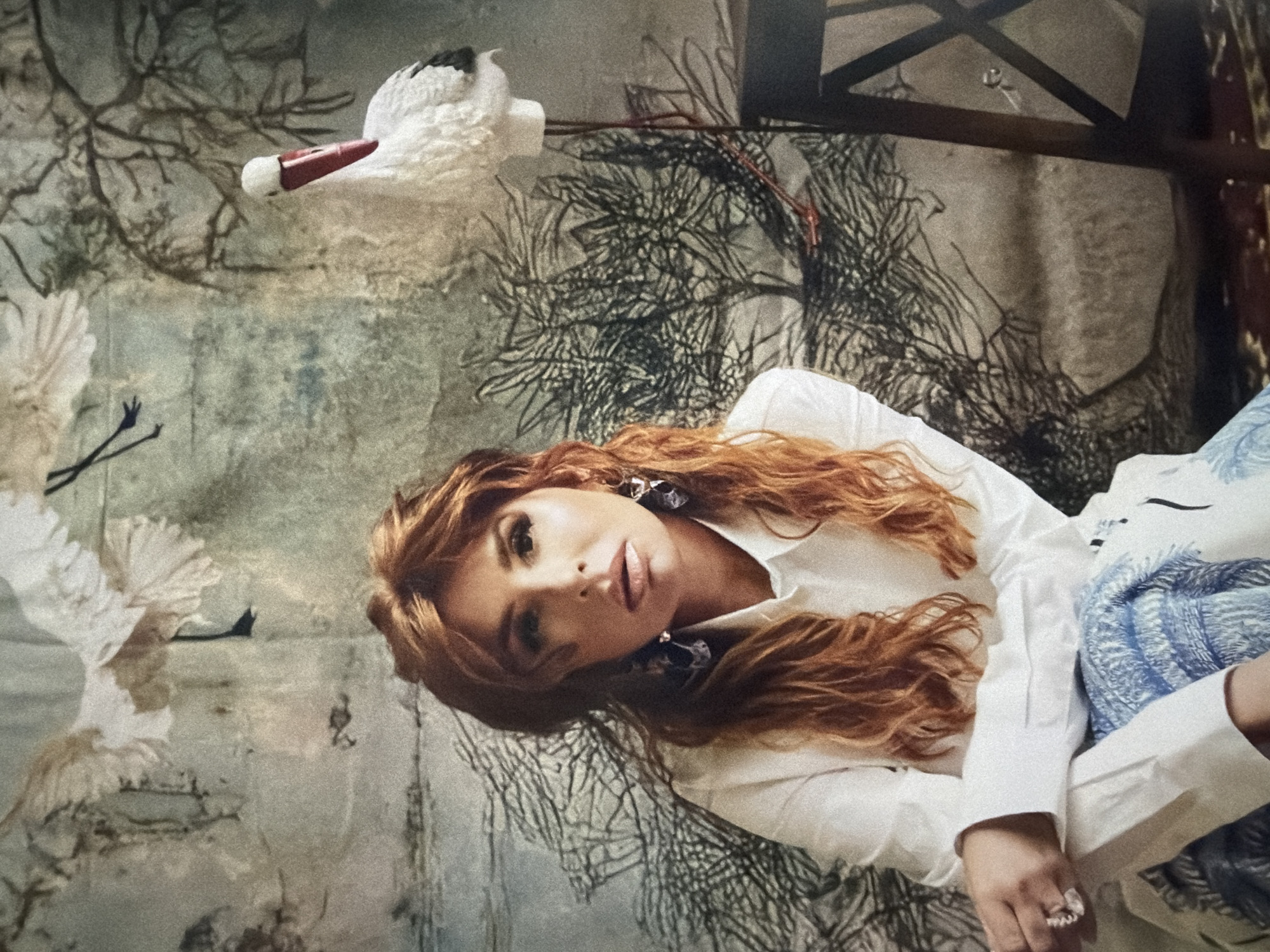 A woman with red hair is lying down in front of a patterned backdrop. The backdrop appears to depict a scene with cranes and branches. The woman is wearing a white shirt, large earrings, and a ring. The overall scene is not obviously identifiable as Moscow, Russia, but the image could be associated with that location.