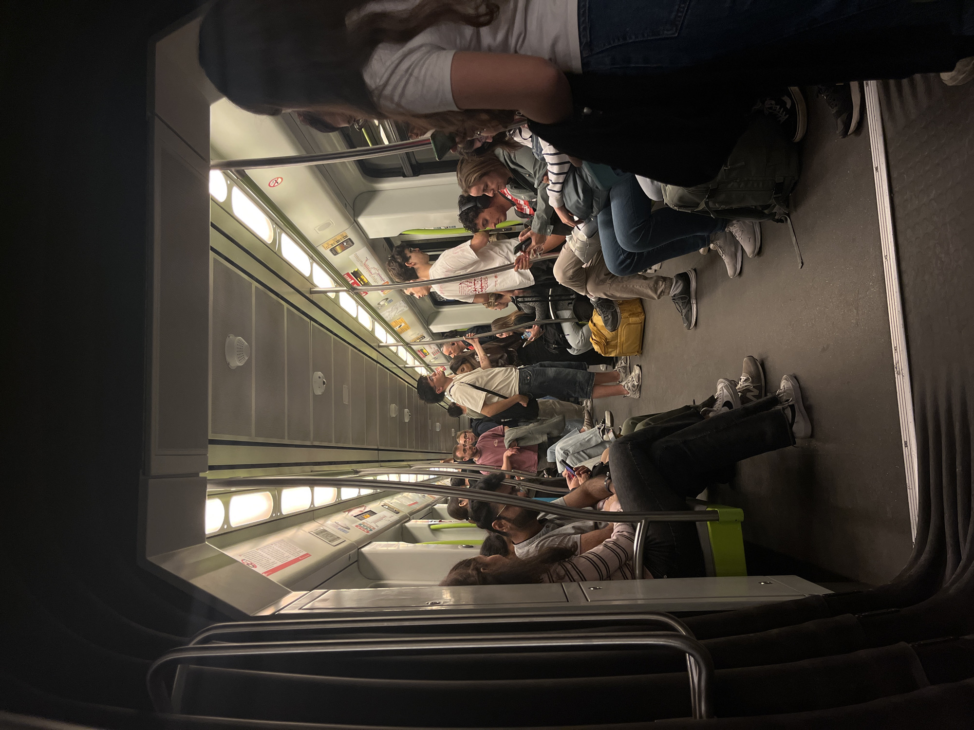 The image captures the interior of a metro train carriage, seen from an elevated perspective looking down the length of the car. The train is moderately crowded with numerous passengers, appearing to be commuters. Most people are seated along the side benches, while some are standing and holding onto the horizontal handrails that run the length of the carriage. The car is brightly lit by overhead fluorescent lights, reflecting off the white and grey interior panels and metallic fixtures.
Passengers are engaged in typical public transport activities; many are looking at their smartphones, some are conversing, and others are simply resting or observing their surroundings. A variety of ages and genders are represented among the passengers. Personal belongings like backpacks and bags are visible on laps or on the floor.
Notable details include the clean, modern design of the train interior, consistent with an urban metro system. The flooring is a dark grey, and the seats are a light, perhaps off-white, color. One seat has a bright green base. Visible text and symbols include a red circular sign with a diagonal line, commonly indicating a prohibition such as 'no smoking' or 'no eating/drinking'. Other smaller informational signs are present on the walls and panels, though the text is unreadable due to distance and resolution. The overall atmosphere seems calm and unremarkable, typical of daily commuting. The image itself is rotated 90 degrees clockwise. Given the context, this is a metro in Valencia, Spain.