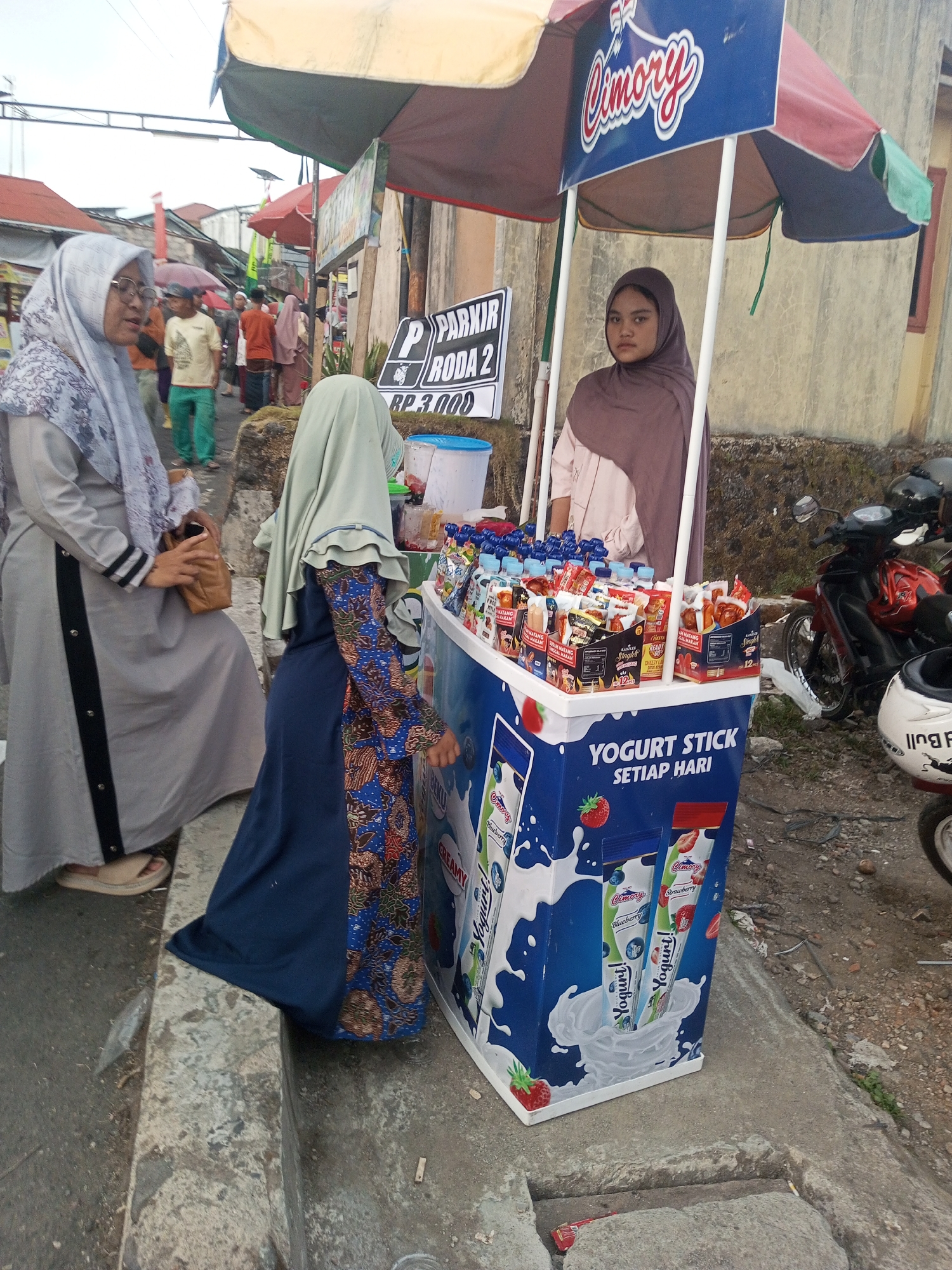 The image shows a street scene in Pandansari, Indonesia. In the foreground, a woman wearing a grey hijab and a long grey dress with black trim is standing near a yogurt stall. To her right, a young woman in a brown hijab stands behind the stall, which is decorated with images of yogurt products and the brand "Cimory". The stall displays various yogurt sticks and bottles. Behind the stall, a red motorcycle is parked. In the background, other people are visible walking along the street, and there are buildings and stalls lining the road, suggesting a marketplace or a busy street. The ground is paved with concrete and asphalt. The overall scene depicts a typical street view in a residential or commercial area of Indonesia.