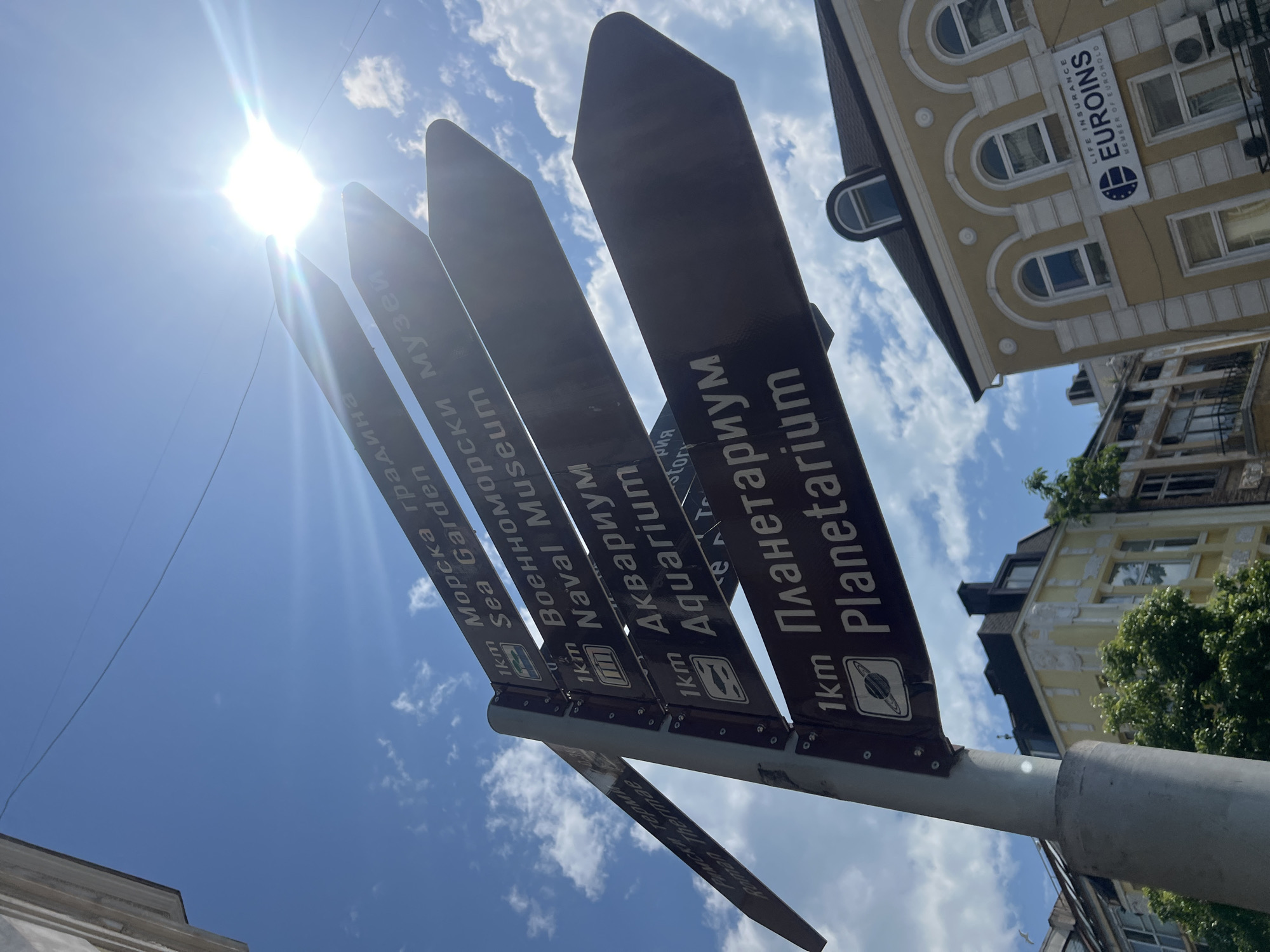 The image captures an upward view of a multi-directional signpost against a bright blue sky with scattered white clouds. The sun is prominently featured in the upper left, casting a strong lens flare across the sky.
The main subject is a dark-colored signpost with multiple arms, each pointing to a different attraction. The text on the signs is in white, featuring both Bulgarian (Cyrillic script) and English, along with illustrative icons. Visible signs indicate directions to:
- "1km Морска градина" / "1km Sea Garden" (with a tree icon)
- "1km Военноморски музей" / "1km Naval Museum" (with an anchor icon)
- "1km Аквариум" / "1km Aquarium" (with a fish icon)
- "1km Планетариум" / "1km Planetarium" (with a planet icon)
The OCR also identifies "Римски терми" / "Roman Thermae" as another destination, likely on a sign further to the left.
In the background, on the right side of the frame, parts of urban buildings are visible. One prominent building is light yellow with intricate white architectural details, including arched windows and moldings. A commercial sign on this building reads "LIFE INSURANCE", "EUROINS", and "MEMBER OF EUROHOLD". Some green tree foliage is also visible near the buildings.
The scene depicts a bright, sunny day in what appears to be a tourist-friendly area of Varna, Bulgaria, indicated by the multi-lingual directional signs to various attractions. No people are visible in the photograph.