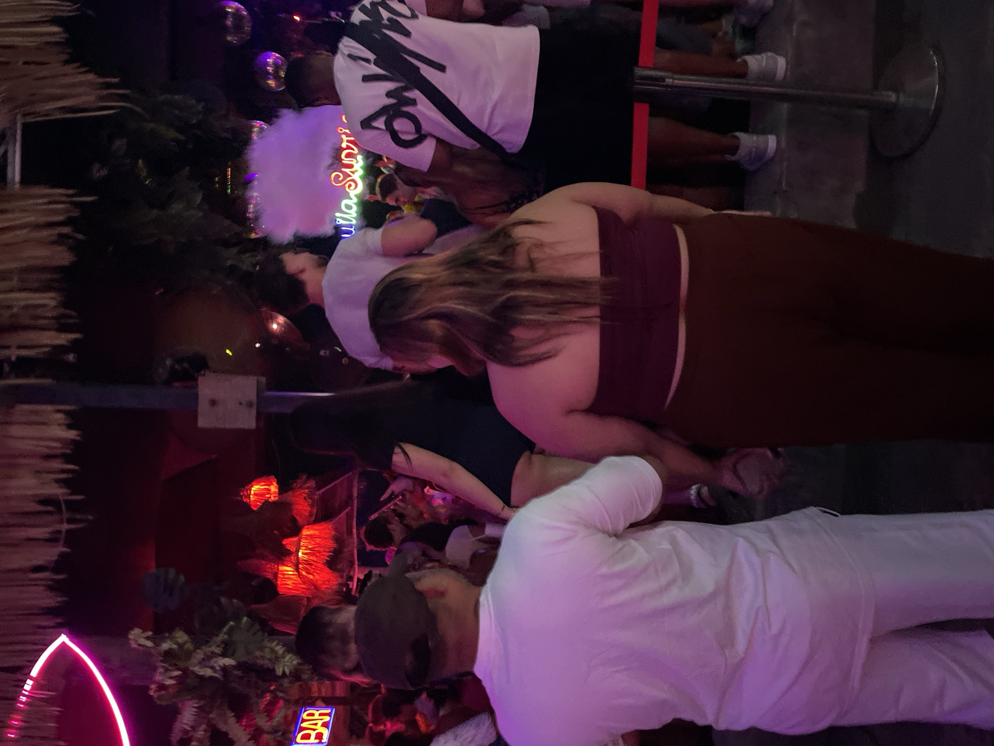 The image captures a nighttime scene at a crowded venue, possibly a bar or club, characterized by vibrant neon lighting and a lively atmosphere. In the foreground, a man wearing a white t-shirt and a brown baseball cap is seen from behind, facing away from the camera. To his right, a woman in a burgundy tube top and brown pants is also viewed from behind, her long brown hair cascading down her back. Behind them, other individuals are visible, some dressed in white t-shirts, and a neon sign that appears to say "BAR" is illuminated on the left side of the frame. Further in the background, a sign with neon lettering that reads "quila Sunri" is visible, accompanied by a cloud of purple smoke or fog. Disco balls are also present, reflecting the colorful lights of the venue. The overall environment suggests a social gathering with music and entertainment.