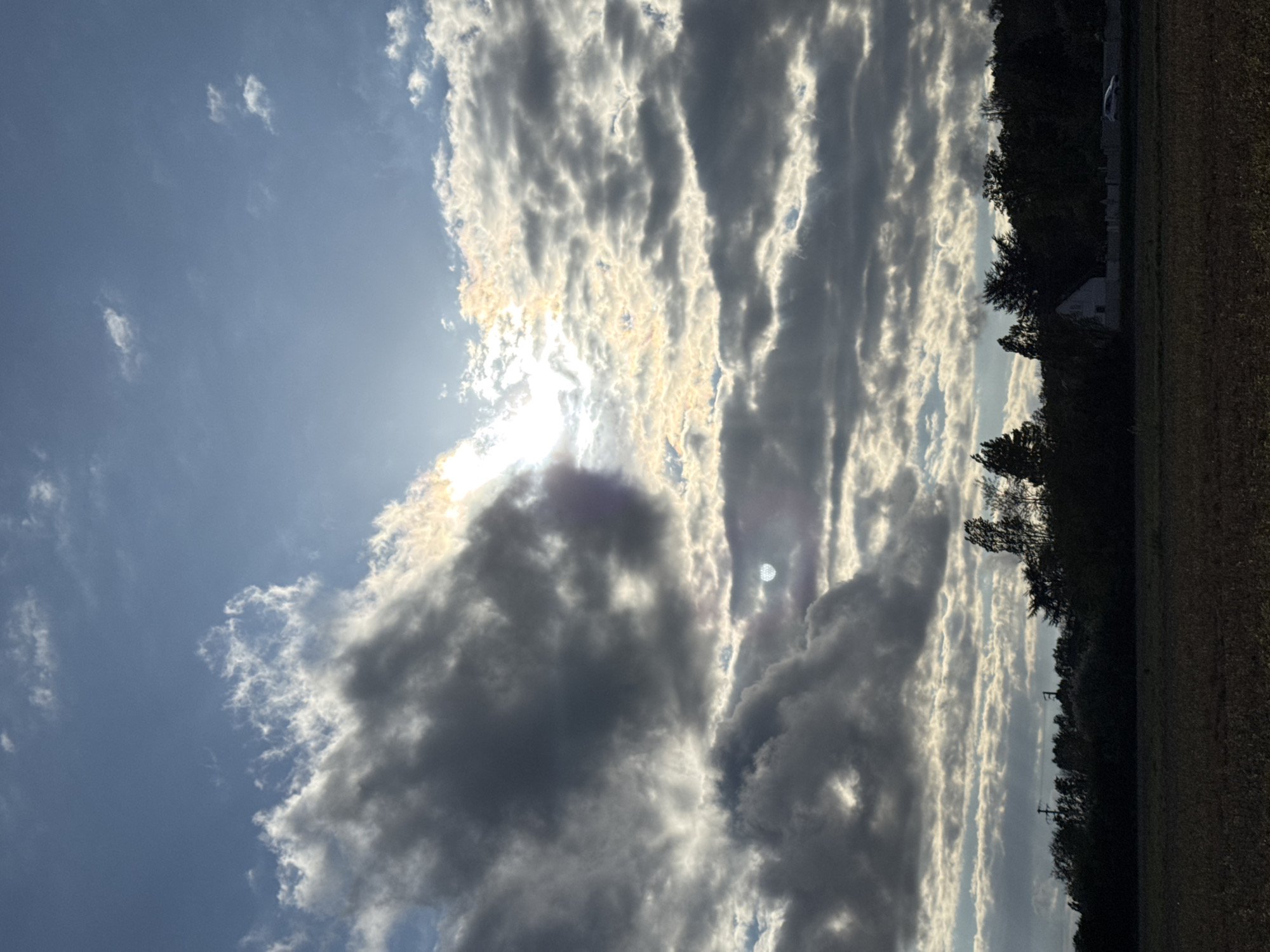 The image shows a daytime sky above Marcoussis, France. The sun is partially obscured by a large mass of striated gray clouds, some with lighter, illuminated edges. A field with low vegetation is visible in the lower right of the frame, bordered by a line of trees. A building is visible amongst the trees. A small patch of blue sky is visible to the left of the clouds.