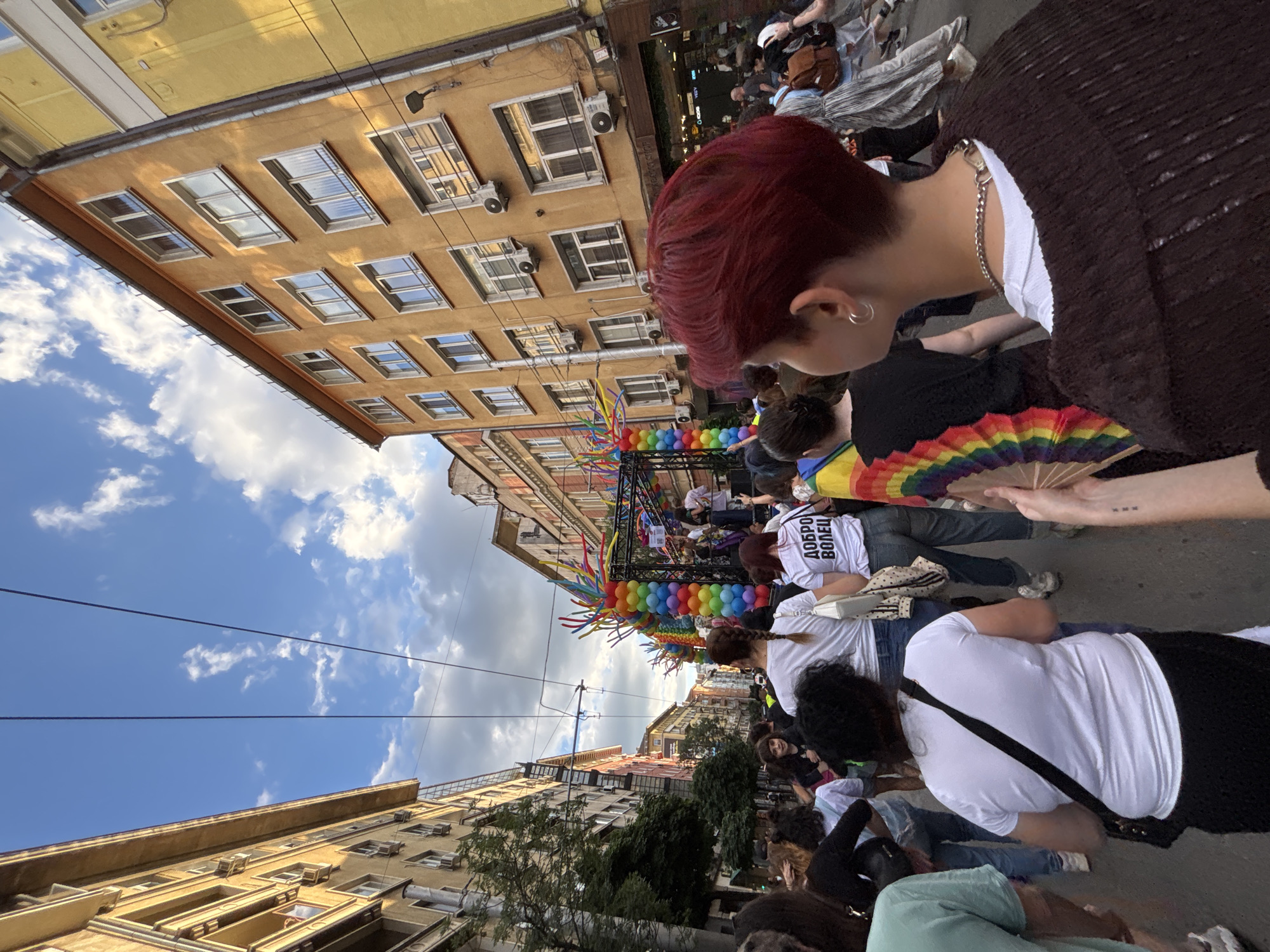 The image depicts a street scene in Sofia, Bulgaria, at a Pride event. The perspective is from within a crowd, looking forward towards a parade float adorned with rainbow-colored balloons. The street is lined with multi-story buildings, typical of a European city, with balconies and air conditioning units visible. The sky is partly cloudy with a bright blue expanse. People are walking in the street, many facing forward, suggesting movement with the parade. Some individuals are holding rainbow flags and fans, common symbols of LGBTQ+ pride. In the foreground, a person with short, red hair is looking towards the parade. The overall atmosphere appears festive and communal.