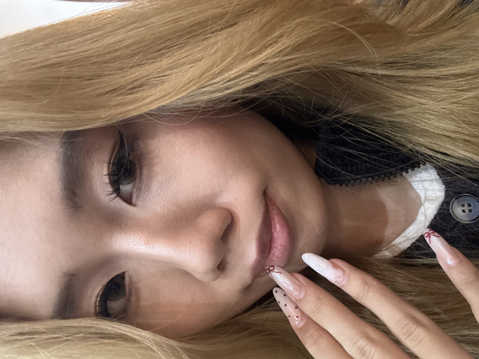 A woman with blonde hair gazes at the camera, her face tilted. She has a fair complexion, dark eyes, and her lips are lightly colored. Her right hand is visible, displaying long, decorated nails. Some nails have red bows, while another has small black dots. The woman appears to be wearing a dark-colored, buttoned sweater with a white collar peeking through. The background is blurred, focusing attention on her face and hand.
