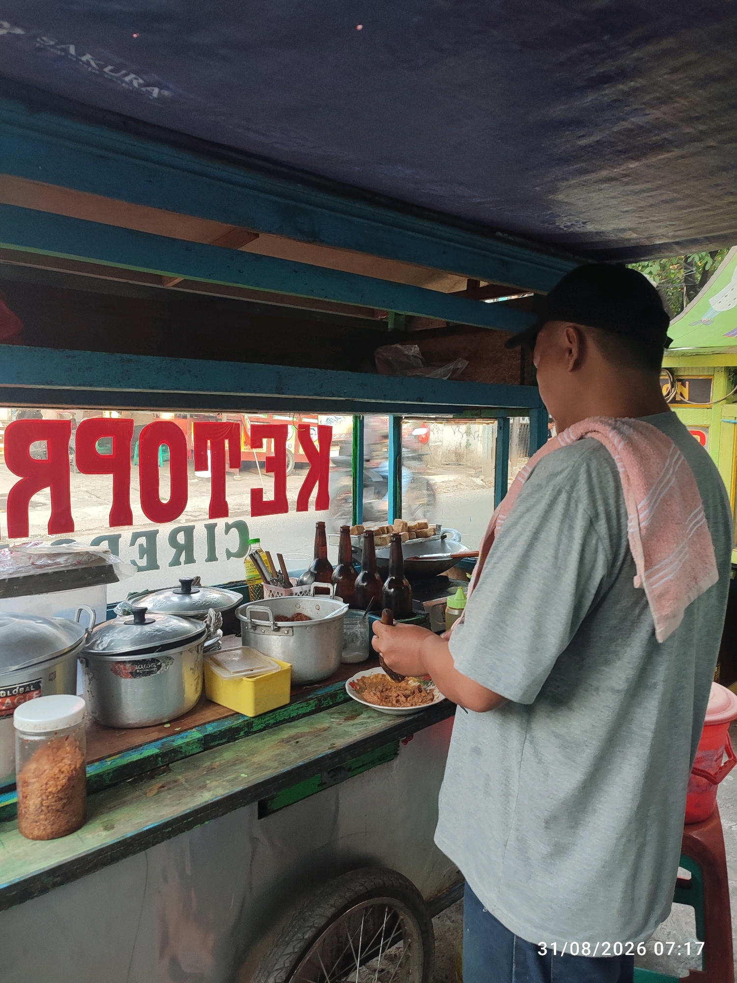 In the city of East Jakarta, Indonesia, a street food vendor is seen preparing a meal from his cart. The cart is covered by a dark blue tarp with the word "SAKURA" visible on the front edge. A man, wearing a grey t-shirt and a black baseball cap, is facing away from the camera, engaged in food preparation. A light pink towel with white stripes is draped over his right shoulder.
The cart's serving window displays a sign in red lettering that reads "KETOPRAK CIREBON". Inside the cart, various food preparation items are visible, including several large aluminum pots with lids, a small yellow container, a glass jar filled with chili flakes, and a plate of food. To the right, there are three brown glass bottles and a white basket containing utensils. The background of the serving window shows a blurry outdoor scene with vehicles and buildings. The timestamp "31/08/2026 07:17" is visible in the bottom right corner of the image.