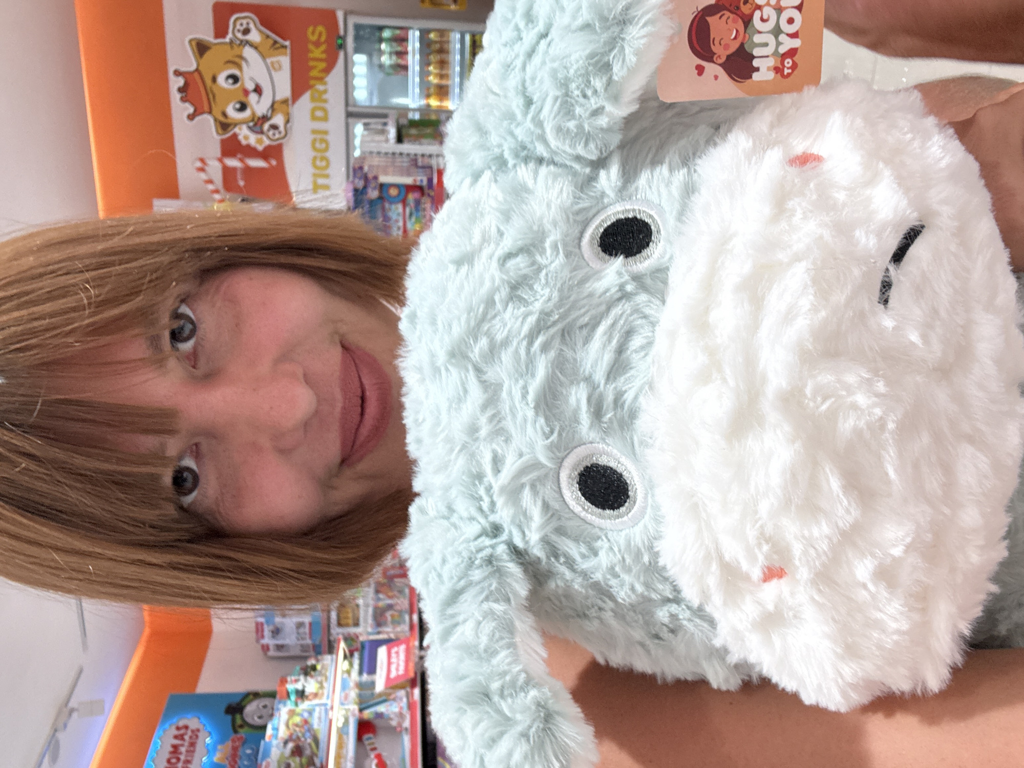 A person with light brown hair is holding a light blue and white plush toy in front of them. The toy has black eyes and a white face. The background appears to be a store aisle, with shelves containing books and toys. There are also drinks visible in a cooler behind the plush toy. The location is Kuta, Indonesia, indicated by the presence of Tiggi Drinks signage, a brand commonly found in that area.