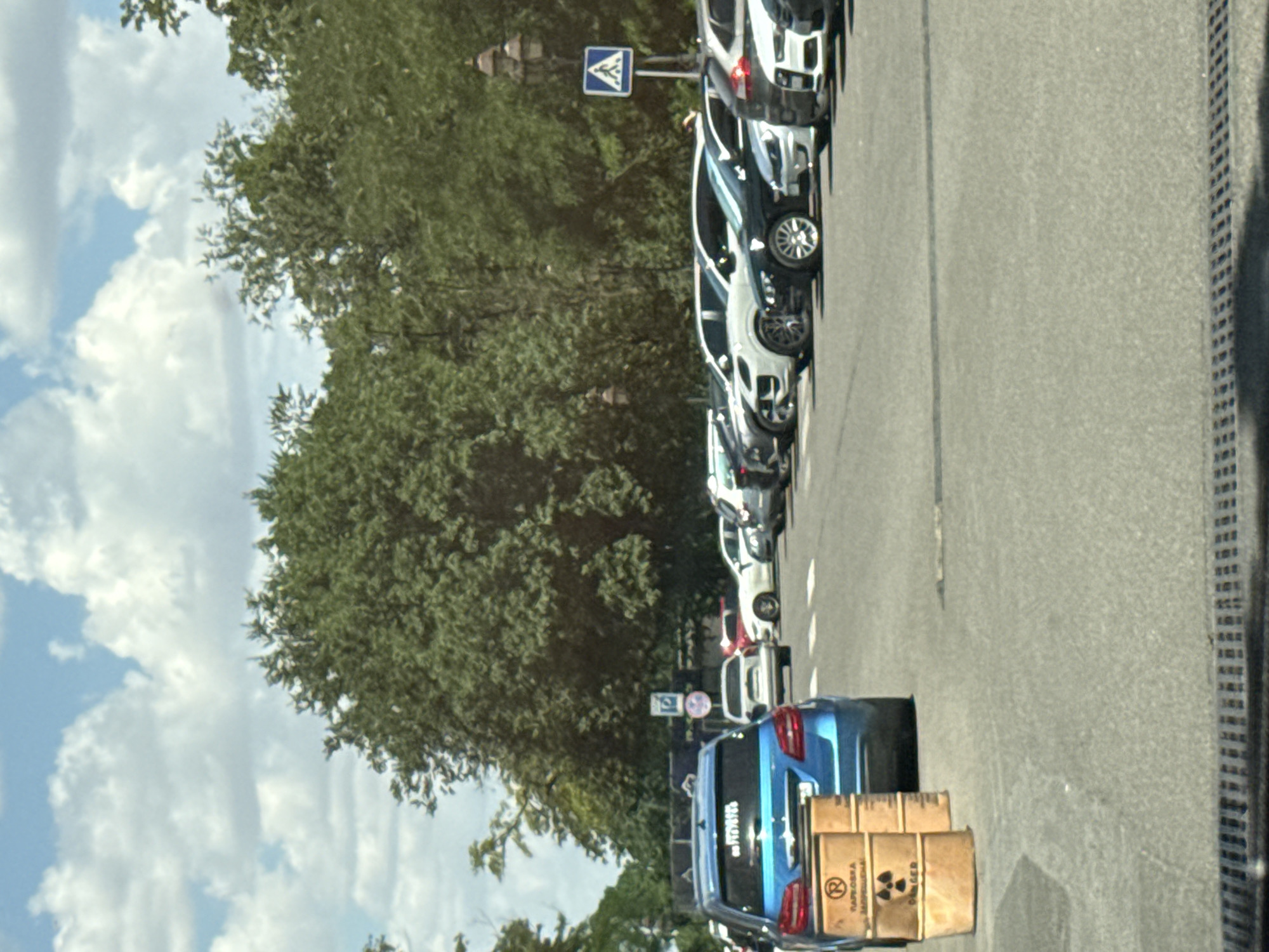 The image shows a parking lot with a row of cars. In the foreground, a blue car is parked, with two barrels next to it. The barrels are made of wood and have a yellow and black hazard symbol, indicating radioactive material. There is text on the barrels in Cyrillic script, which translates to "RADIOACTIVE MATERIAL" and "DANGER". The parking lot is paved, and there is a drain grate in the lower part of the image. In the background, there are trees and a blue sky with white clouds. There is also a street sign visible in the background. The scene appears to be a public area, possibly a street or a parking lot in a city. The presence of radioactive material barrels suggests a potentially hazardous situation. It is not possible to determine the exact location within Odesa, Ukraine from the image alone, but it is a street or parking area in a city setting.