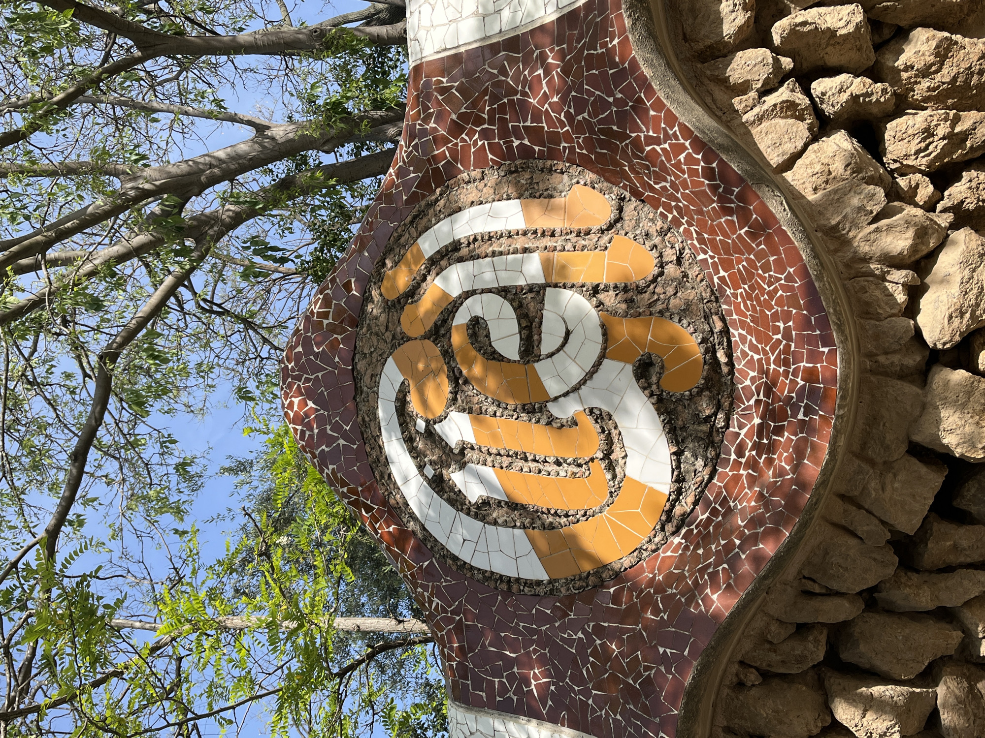This media file captures an outdoor scene featuring a distinctive mosaic-covered structure, likely part of Antoni Gaudí's Park Güell in Barcelona, Spain. The image is composed diagonally, with a vibrant blue sky and a network of tree branches and green leaves occupying the upper left quadrant.
The prominent subject is a segment of a curved wall or architectural element, adorned with colorful broken tile mosaics (trencadís). The main body of this structure is covered in irregular pieces of brown tile. Within this, a circular emblem is embedded, featuring a swirling, organic pattern made of white and orange/yellow mosaic pieces, suggesting an artistic or decorative motif. This central design is bordered by smaller, pebble-like stones.
To the right of the mosaic structure, a natural stone wall constructed from rough-hewn, light-colored rocks is visible, partially in shadow. The sun casts distinct shadows across the mosaic, indicating bright daylight. The sky is clear, and the trees appear to be in full leaf or budding, suggesting spring or summer. No human activity or visible text is present in the image. The unique mosaic style and natural materials are strong indicators of its location.